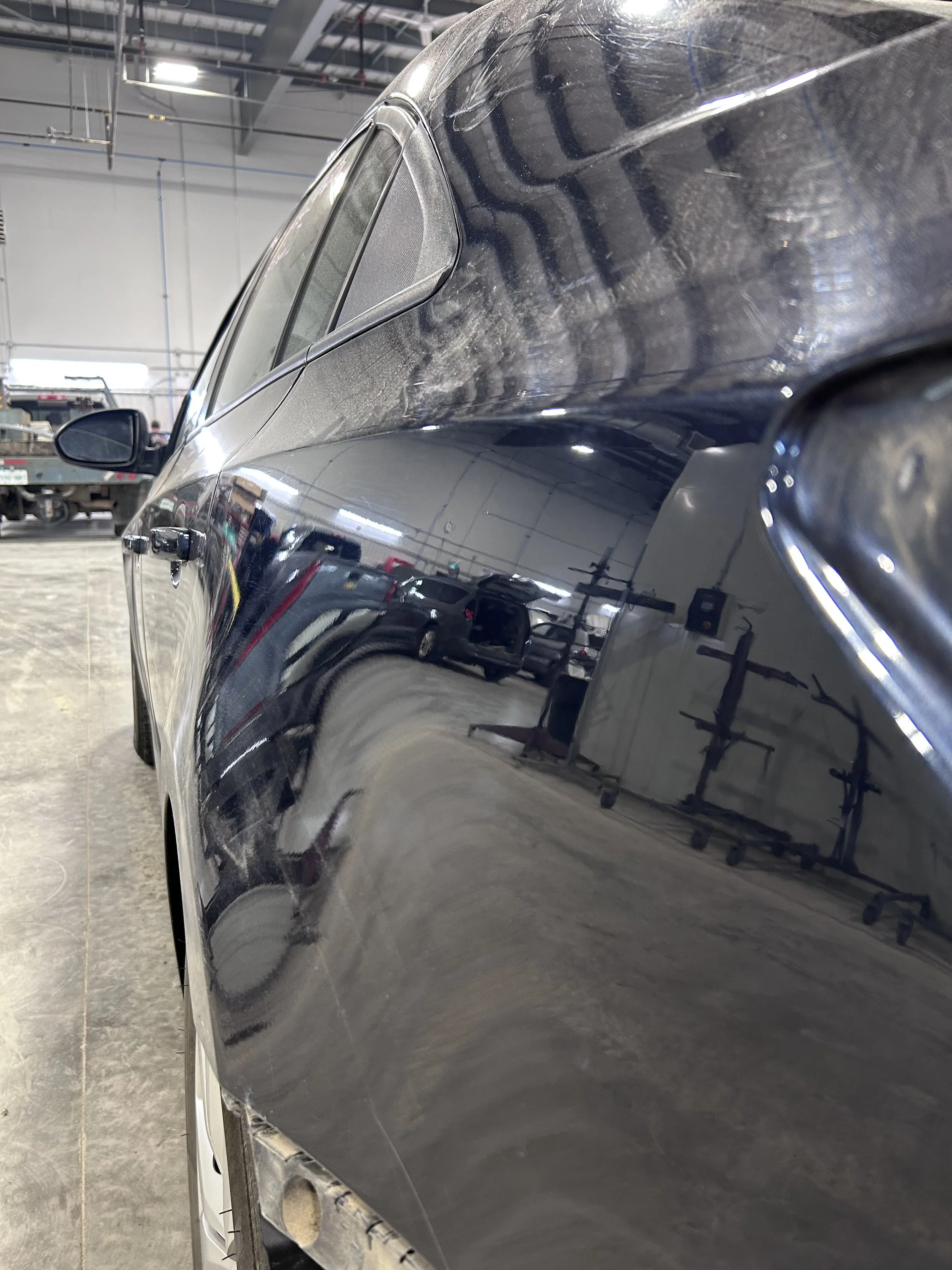 Close-up of a black car's side panel with visible scratches and dirt, inside a garage or workshop with tools and equipment reflected on its surface.