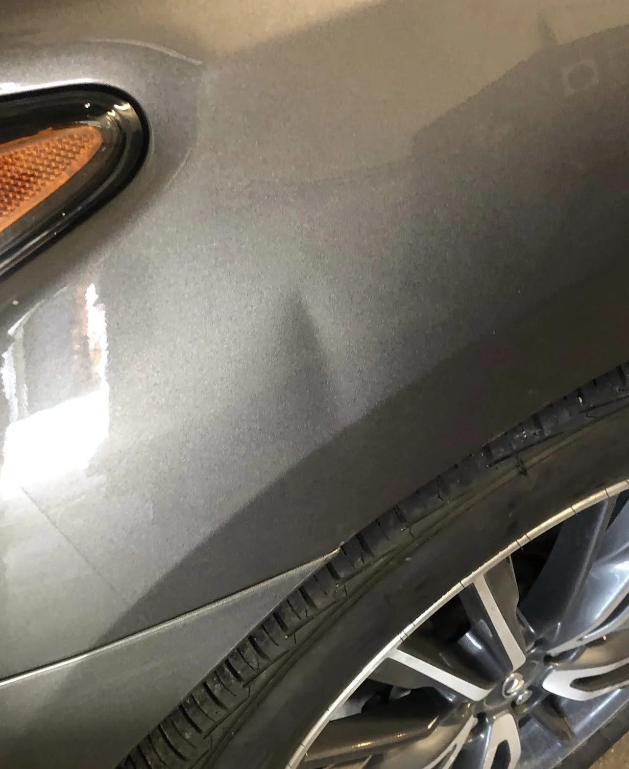 Close-up of a gray car's front fender, part of the headlight, and a portion of the wheel with tire and alloy rim visible.