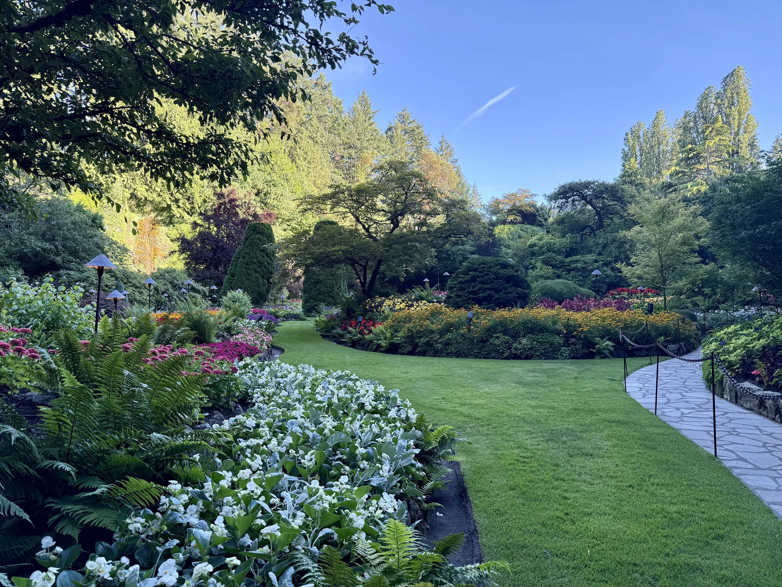 A scenic garden with a curved stone pathway lined with black chains, lush green grass, colorful flower beds, and tall trees under a bright blue sky.