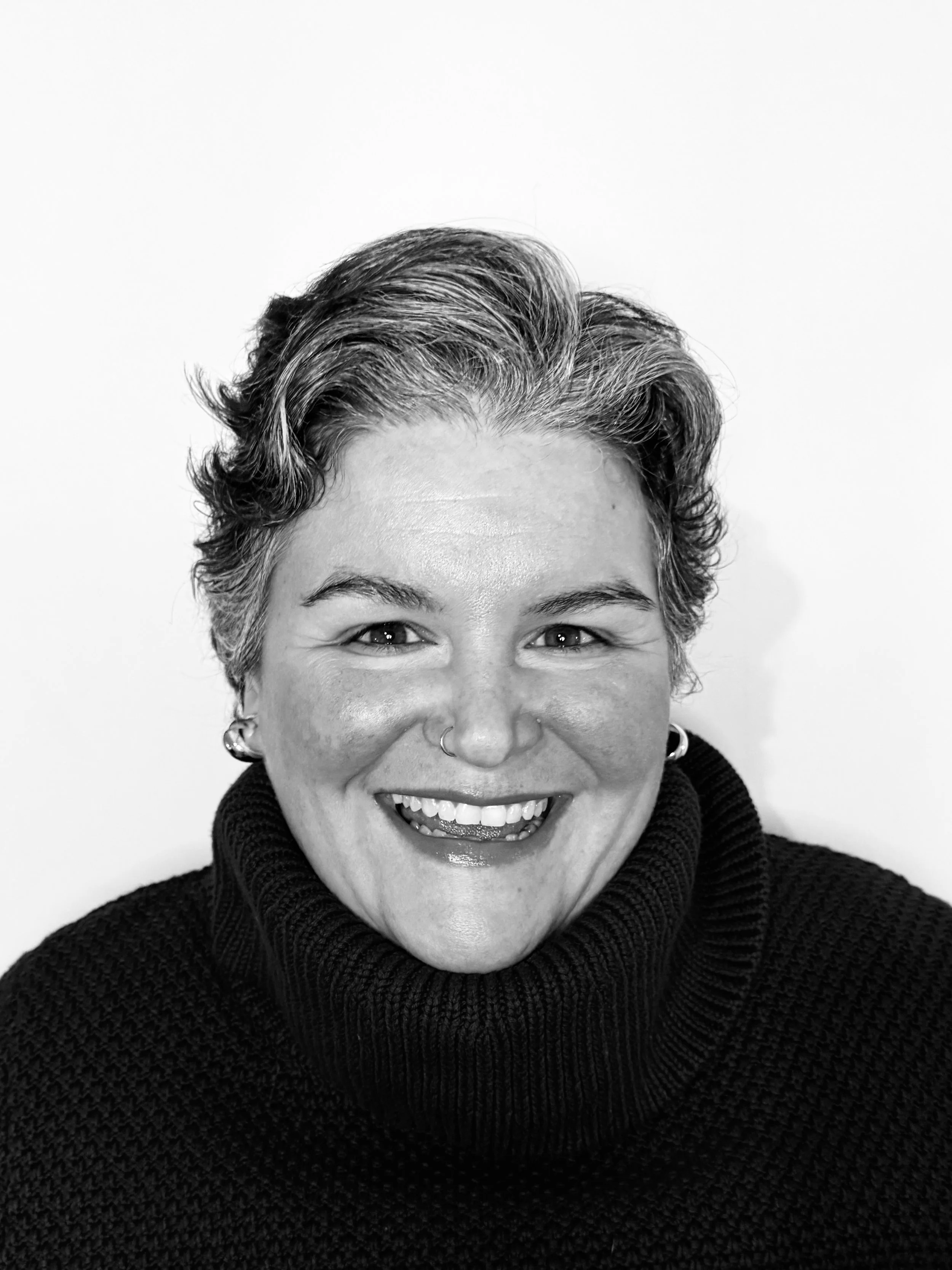 Black and white portrait of a smiling woman with short, wavy gray hair, wearing a dark turtleneck sweater and earrings.
