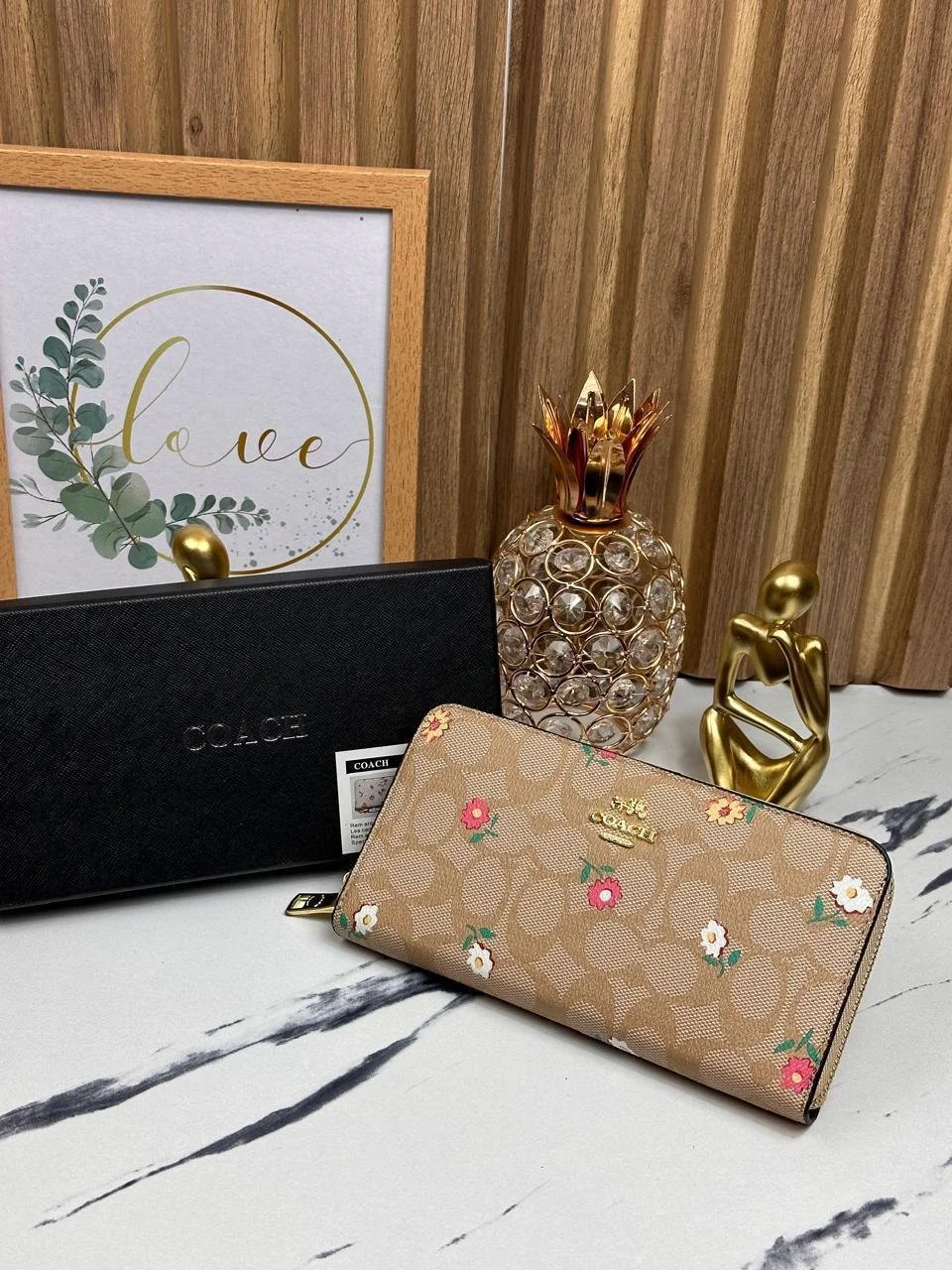 Dos bolsos de marca Coach, uno negro y otro beige con flores, junto a decoraciones doradas y una ilustración con la palabra 'love' enmarcada en una pared de madera.
