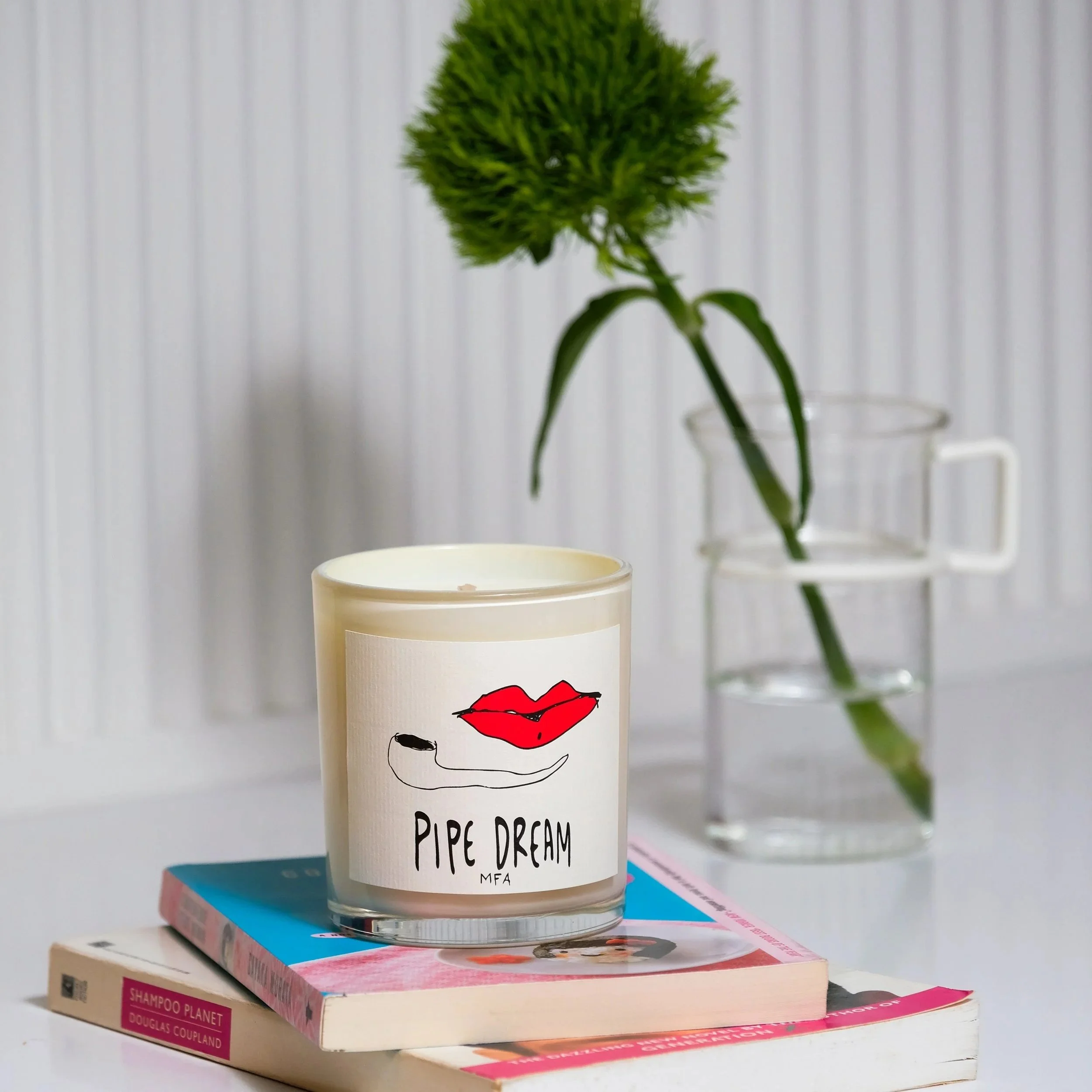 A white candle with a label featuring red lips, a pipe drawing, and the text 'Pipe Dream MFA,' placed on top of two books, with a glass vase holding a green flower in the background.