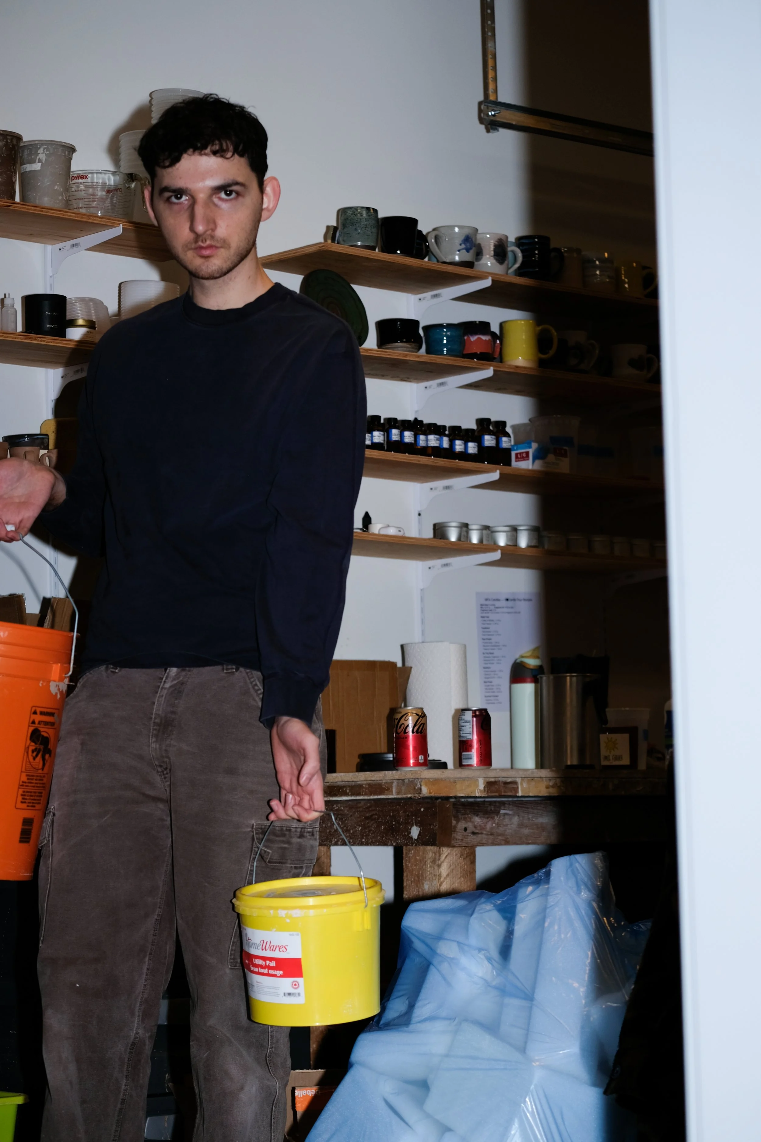Young man holding a yellow bucket inside a storage room with shelves filled with cups, bottles, and other supplies.