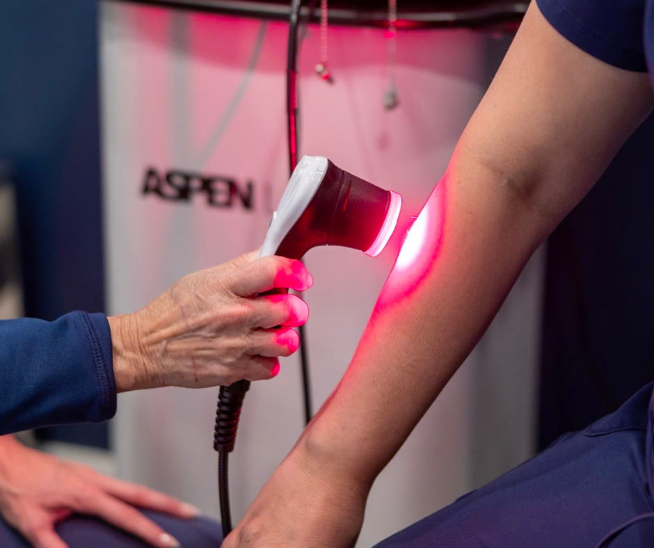 A healthcare professional uses a handheld laser device on a patient's forearm in a clinical setting.