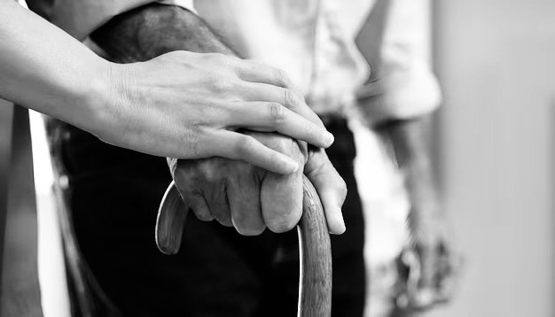 Close-up of a younger person's hand holding an older person's hand gripping a cane, depicting arthritis, carpal tunnel syndrome, tendonitis, or injuries