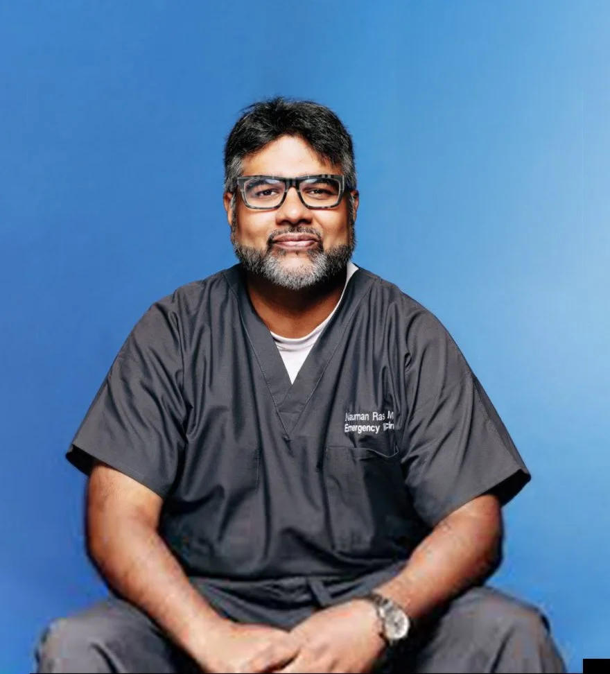 A man with glasses and gray beard wearing black scrubs, sitting against a blue background.