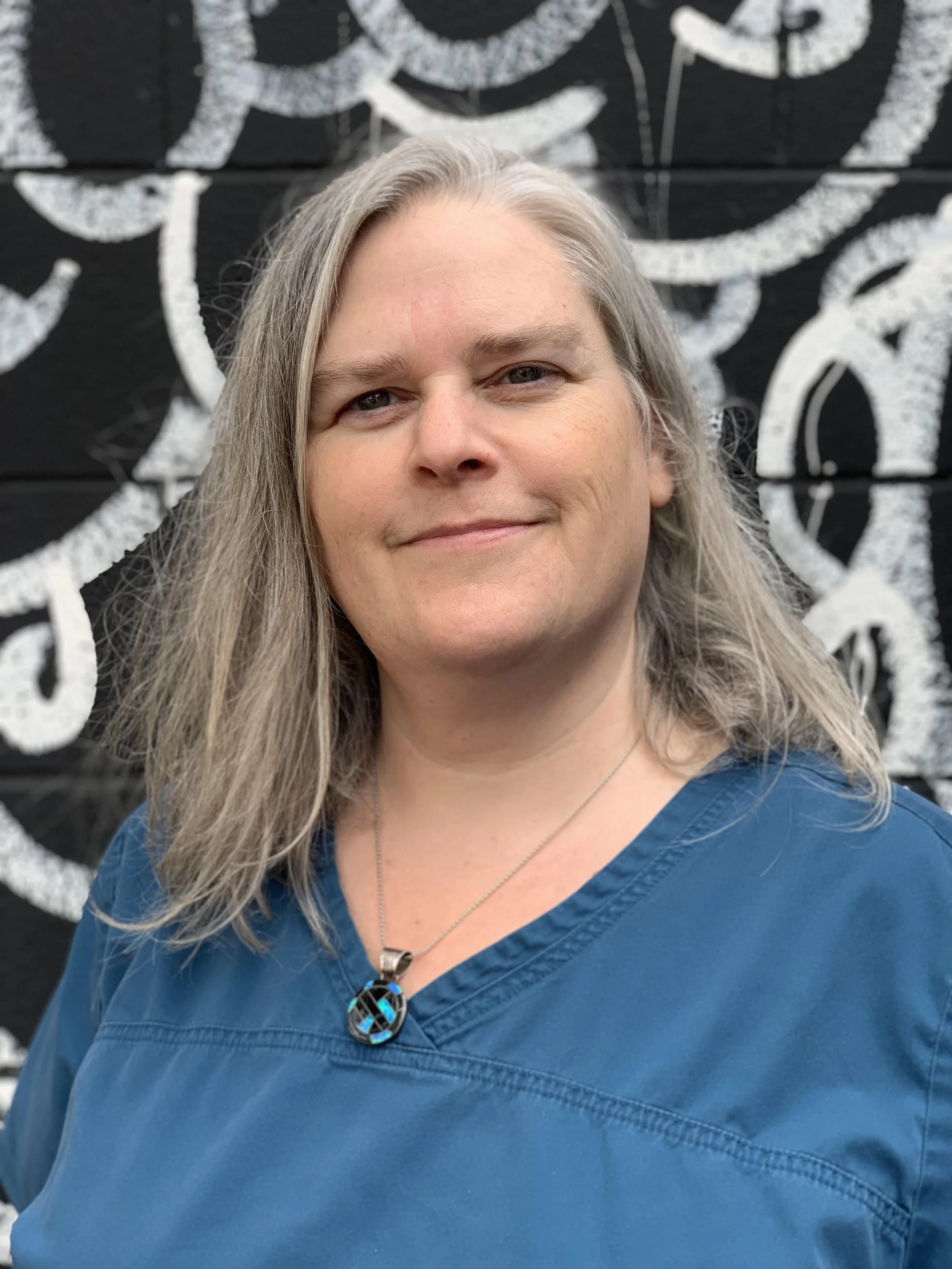 A woman with long gray hair wearing a blue medical scrub top and a peace sign necklace, standing in front of a black wall with white abstract swirls.