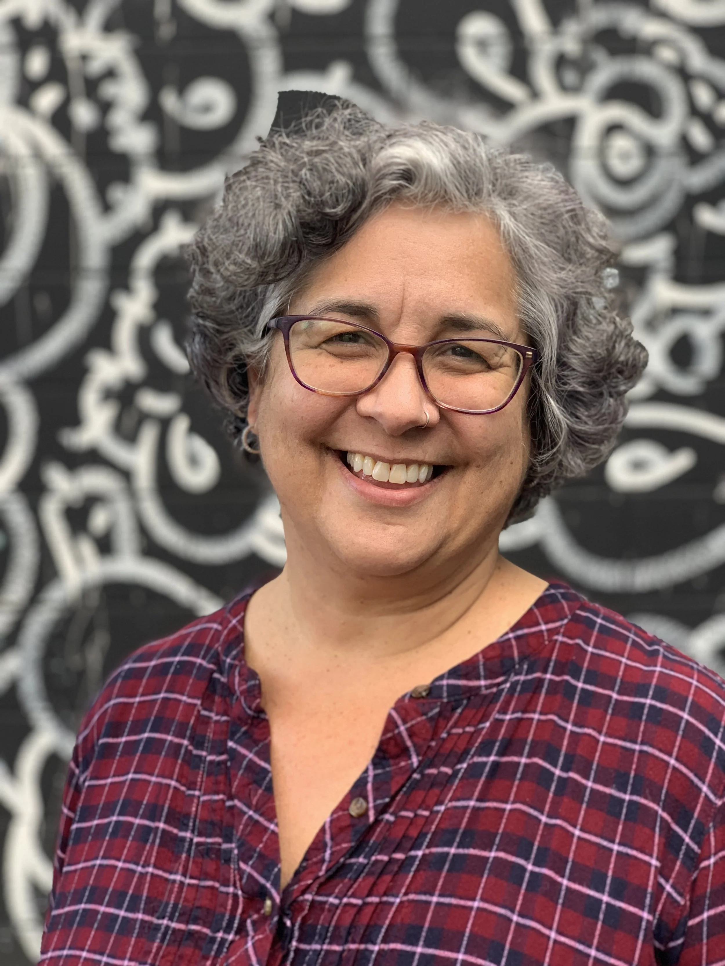 Smiling woman with curly gray hair, glasses, and a nose piercing, wearing a red and blue plaid shirt, standing in front of a black and white illustrated background.