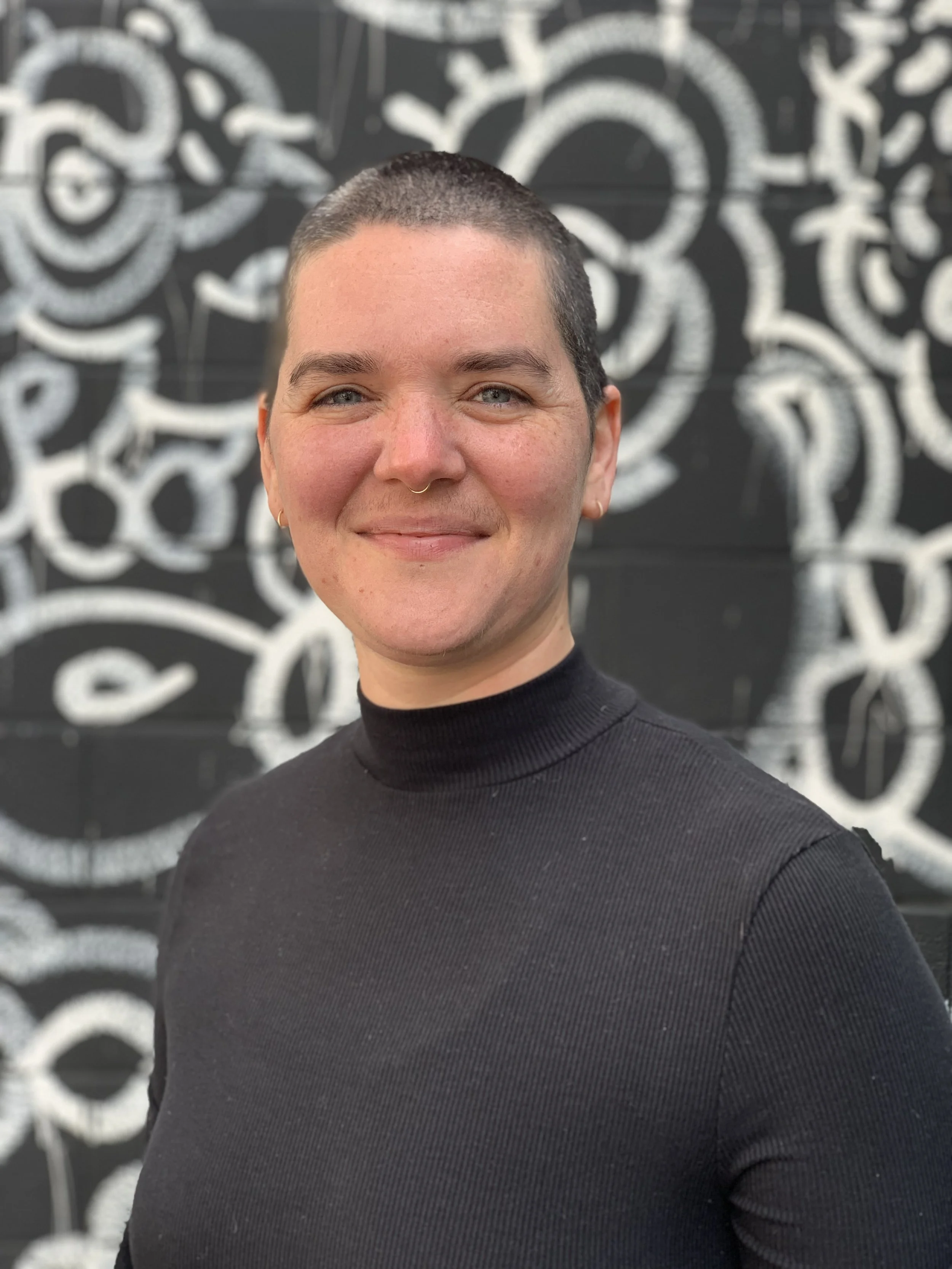 A person with short dark hair, fair skin, blue eyes, wearing a black turtleneck, smiling in front of a black wall with white abstract graffiti design.