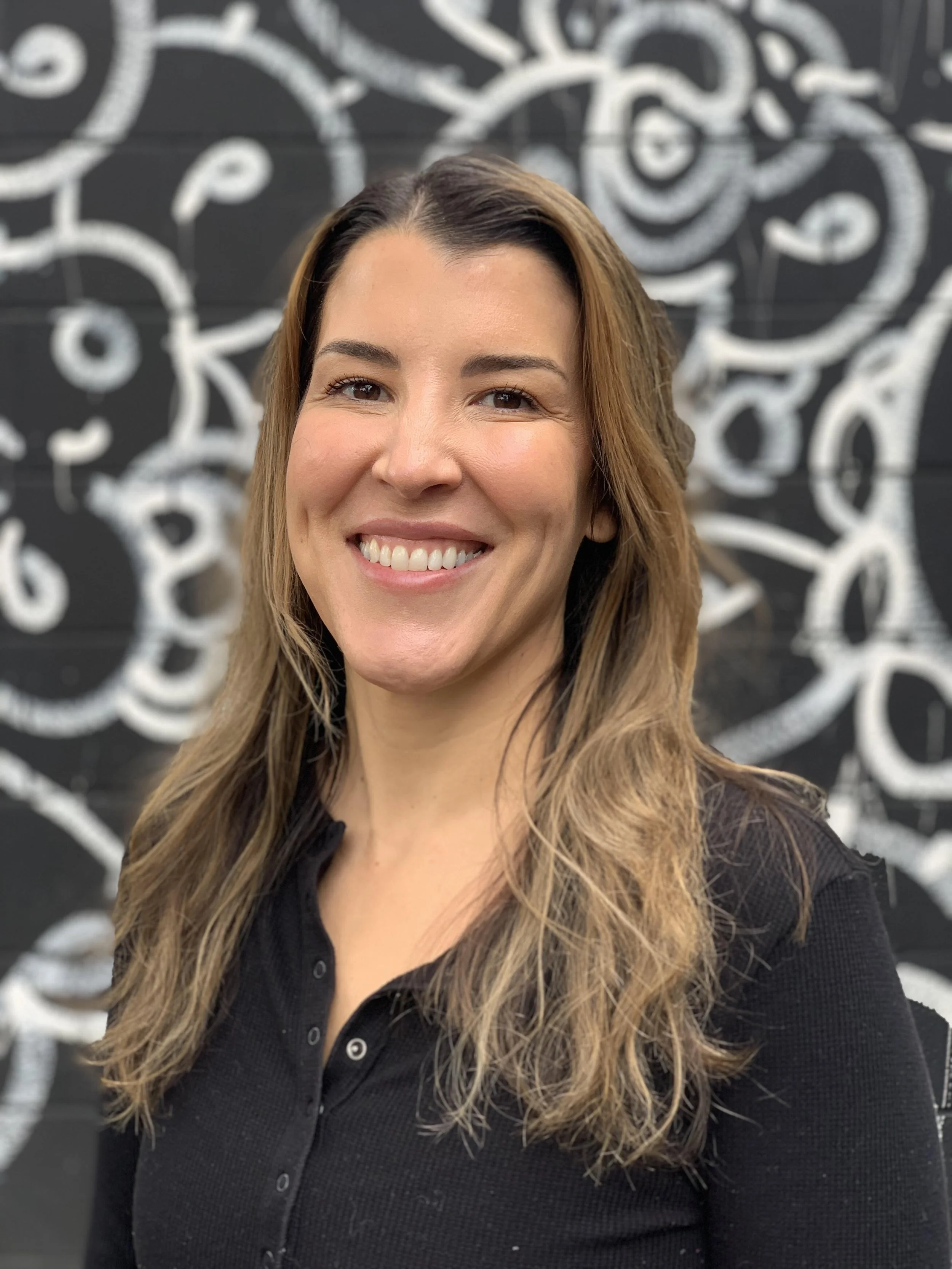 A woman with long brown hair smiling in front of a black and white abstract wall mural.