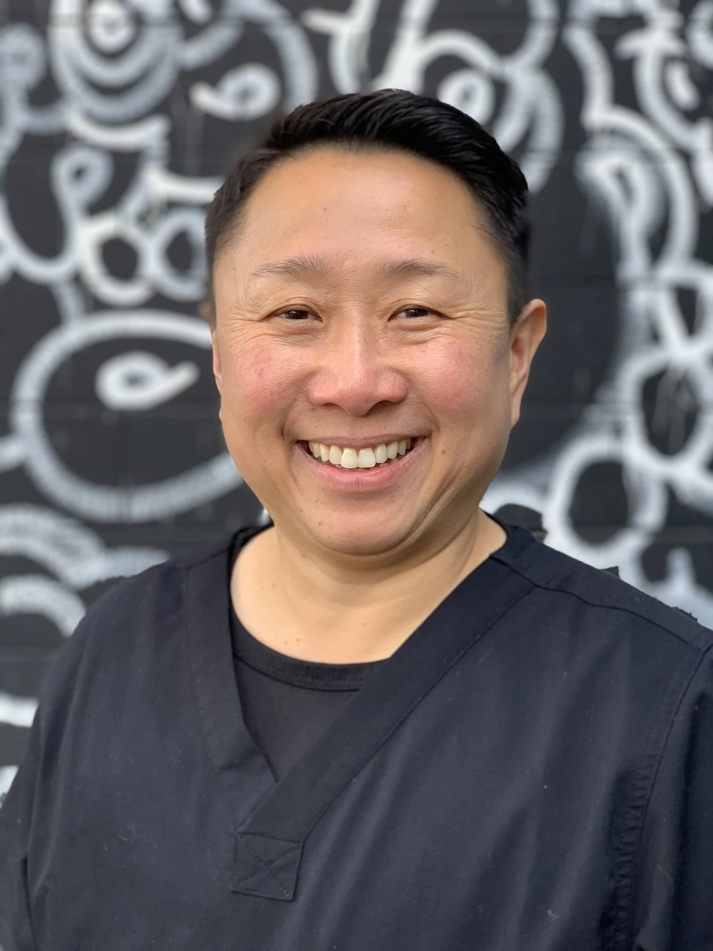 A man smiling in front of a black and white graffiti mural.