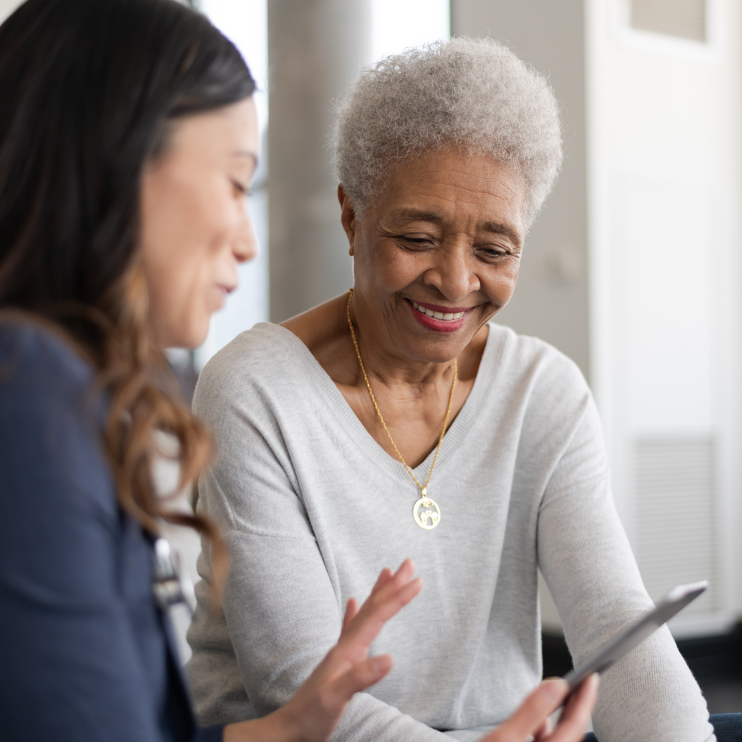 An elderly woman and a young woman sitting together, smiling and looking at a smartphone the young woman is holding.