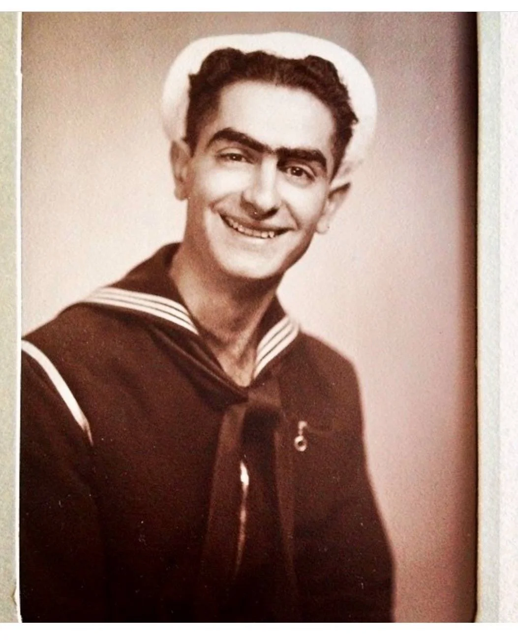 A black-and-white portrait of a young man in a sailor uniform with a cap, smiling at the camera.