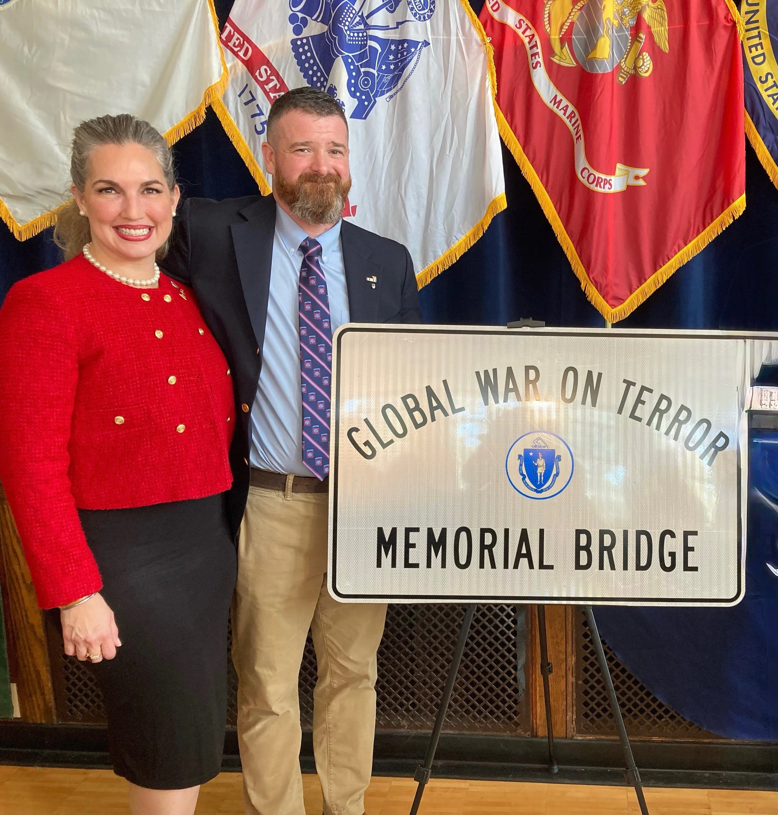 A woman in a red jacket and a man in a dark suit stand beside a sign that reads 'Global War on Terror Memorial Bridge'. Behind them are flags, including the United States Marine Corps flag.