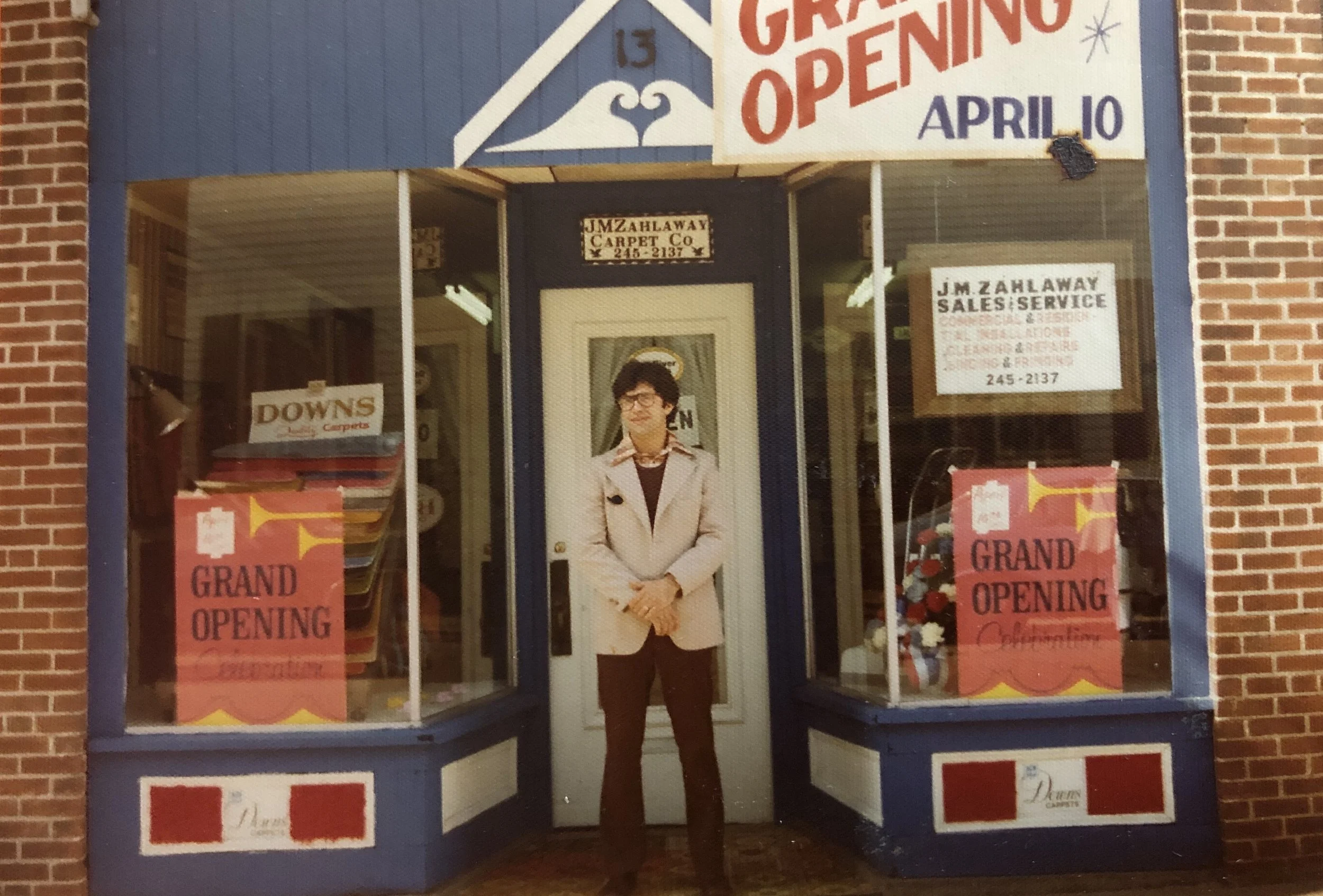 A man is standing in front of a carpet store with a sign indicating a grand opening event. The store has large glass windows displaying signs and carpet samples, and the woman is dressed in a light-colored coat and dark pants, wearing glasses.