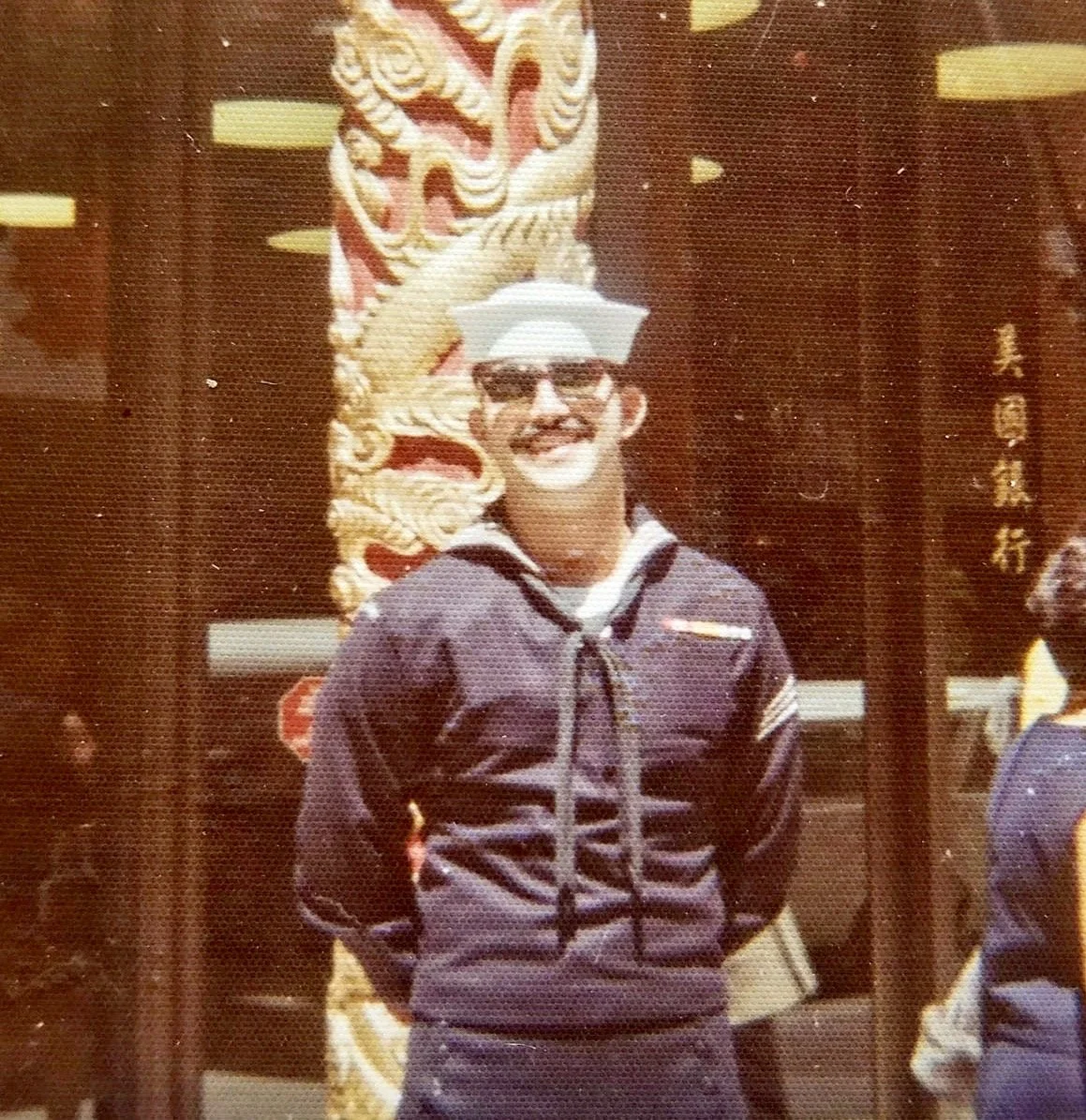 A man wearing sunglasses, a white hat, a dark jacket, and smiling, standing in front of a traditional Asian decorative dragon sculpture.