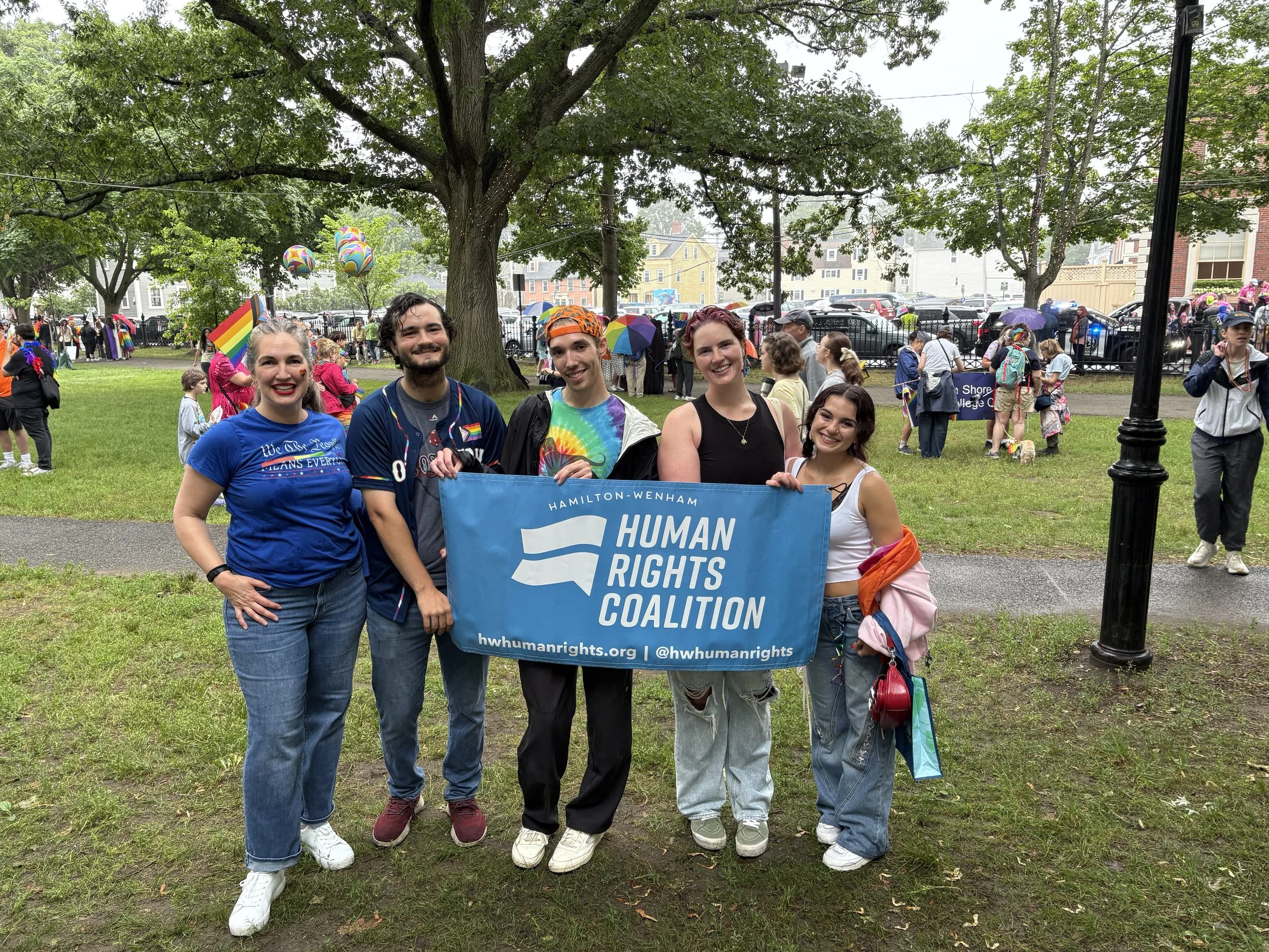 Group of five people standing on grass holding a blue banner that reads 'Hamilton-Wenham Human Rights Coalition.' They are at a pride parade or event, with many other people, flags, and rainbow-themed decorations in the background.