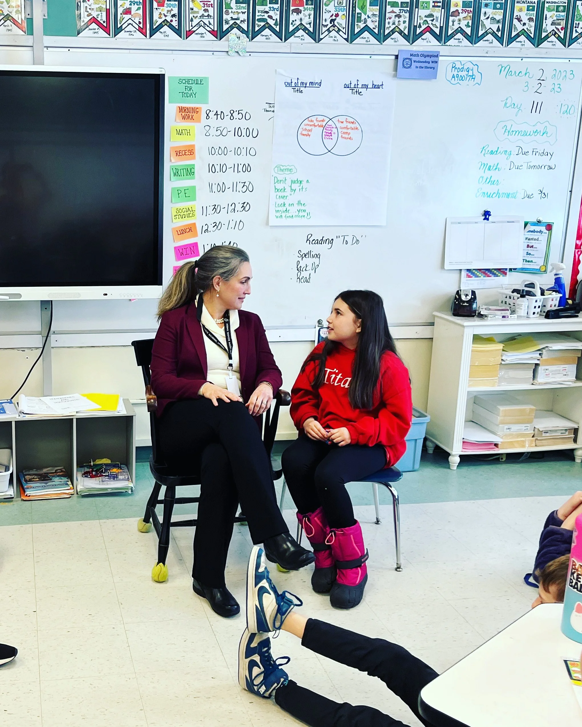 A teacher and a student sitting and talking in a classroom, with a whiteboard behind them filled with schedules, notes, and colorful charts. There are other students seated in the classroom, visible partially.