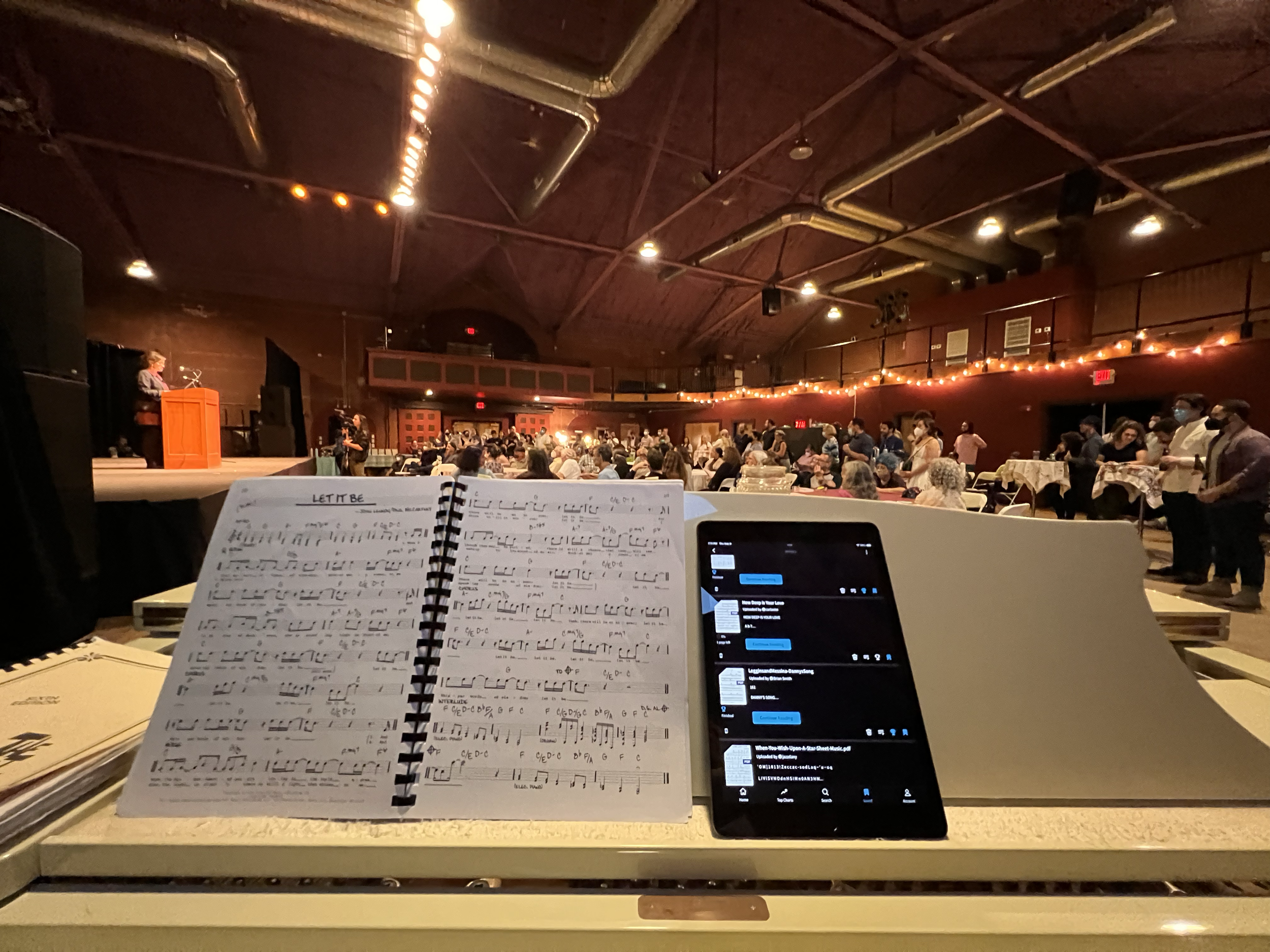 View from the stage at a concert or event showing a music stand with sheet music, a tablet, and an open book in the foreground. In the background, a large audience is gathered in a spacious hall with a high, wooden ceiling, string lights, and a perso