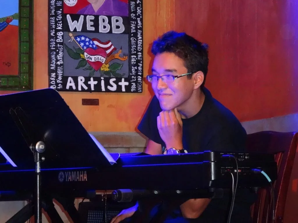 A man with glasses and dark hair playing a black Yamaha keyboard in a room with wooden walls, and a colorful poster of an artist named Webb behind him.