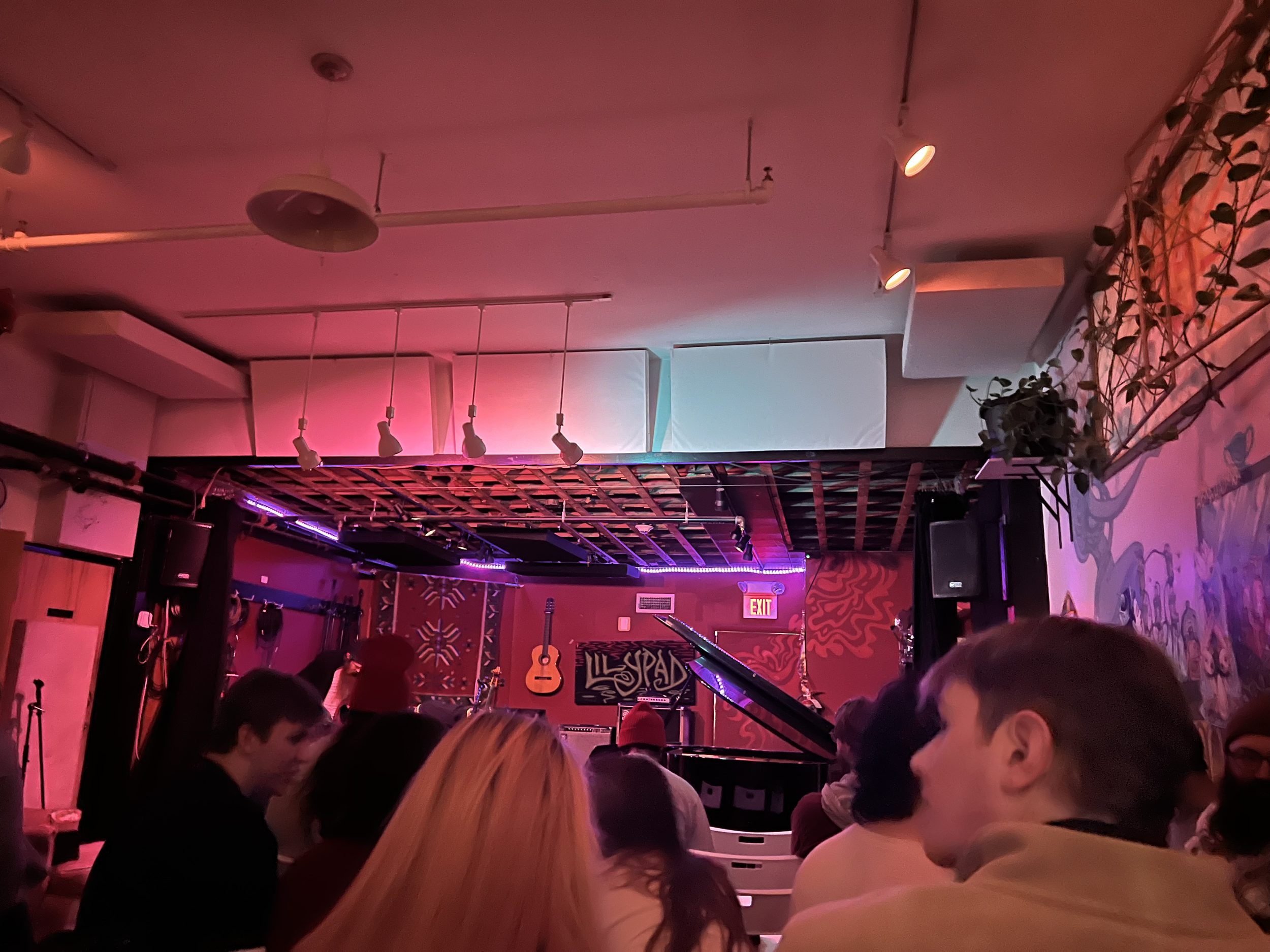 A crowded indoor music venue with people seated, a piano at the front, and a guitar hanging on the back wall. Colorful neon lights illuminate the stage area.