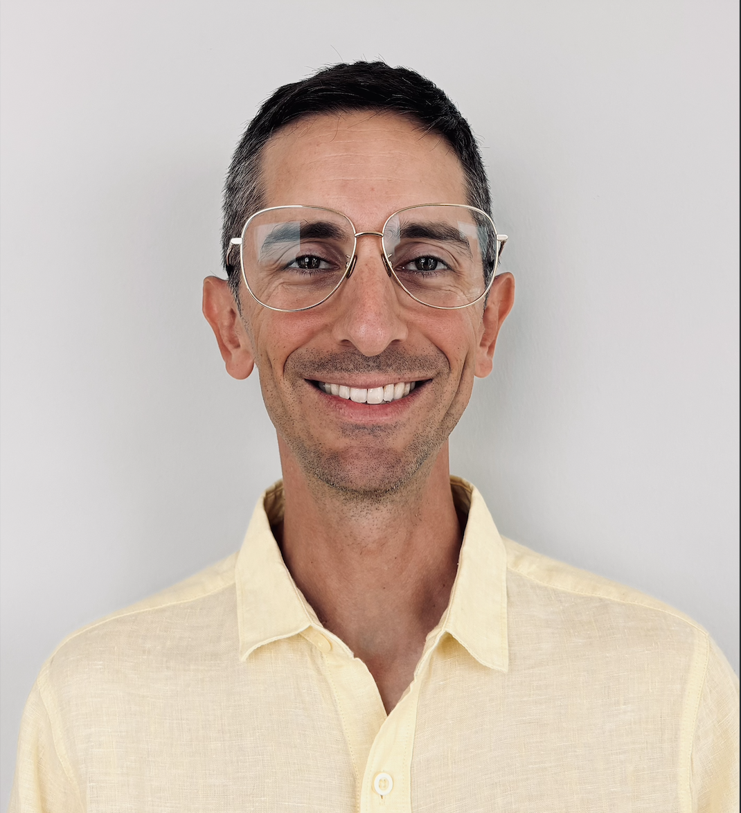 A smiling man with short dark hair, wearing glasses with large round frames and a light yellow collared shirt, standing against a plain white background.