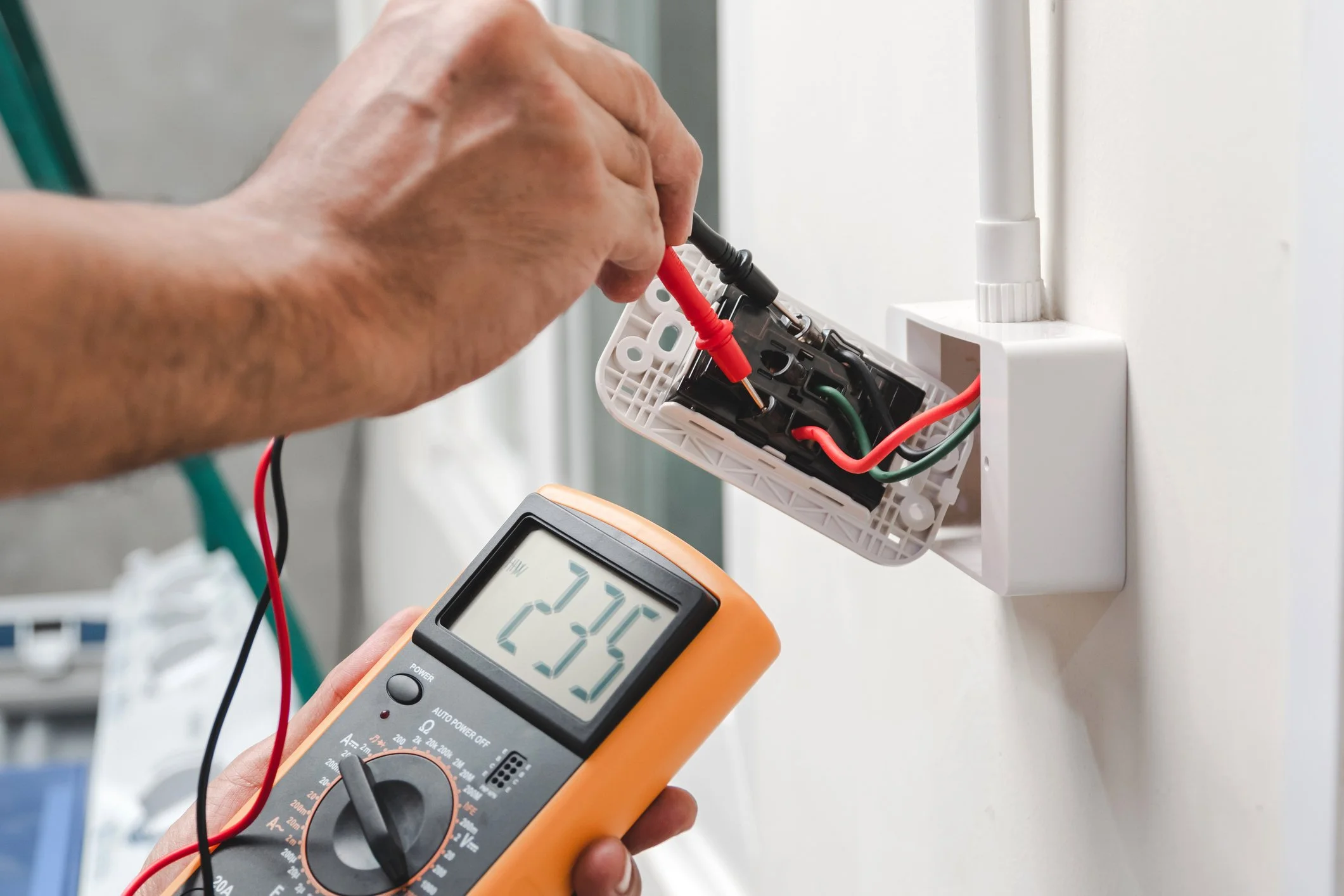 A person using a multimeter to test electrical wiring inside a wall switch box.