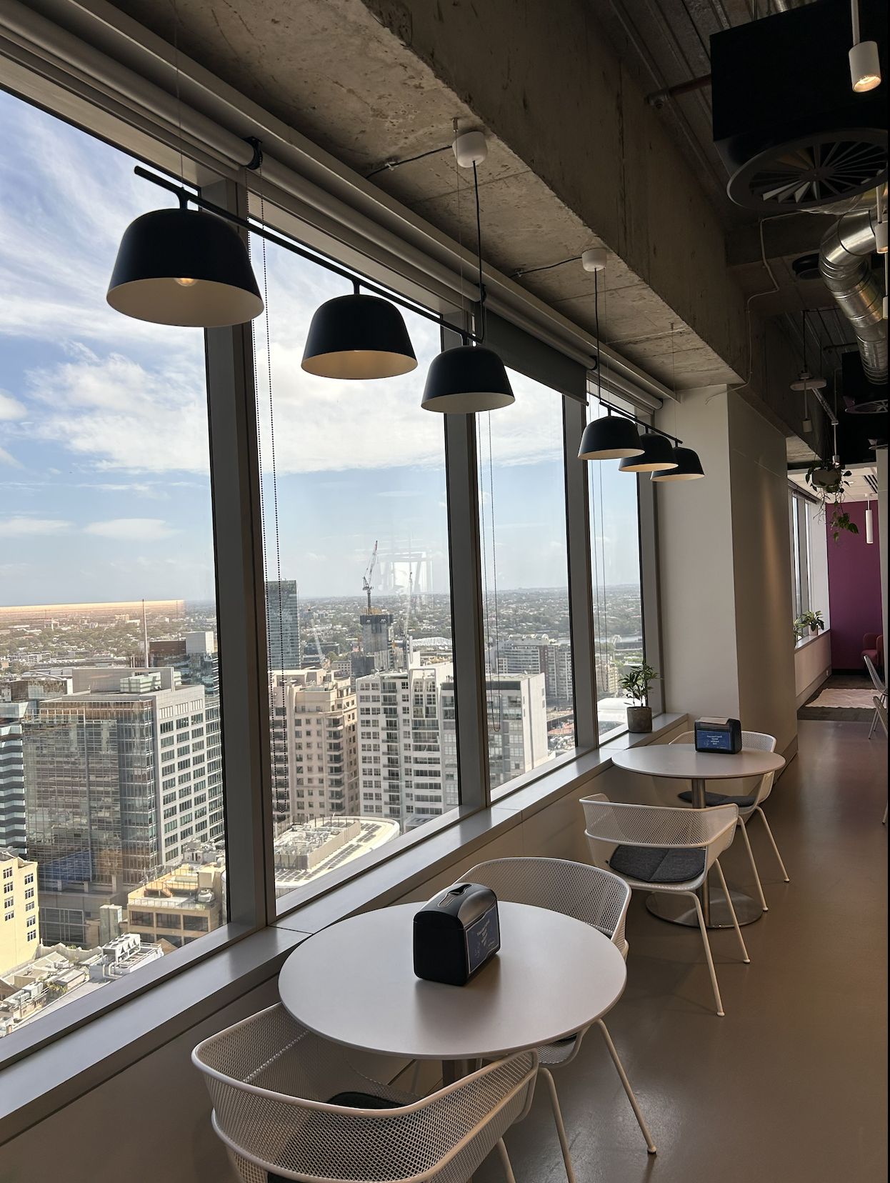 Interior of a high-rise cafe or lounge with white tables and chairs by floor-to-ceiling windows showcasing a cityscape view, hanging black pendant lights, and potted plants.