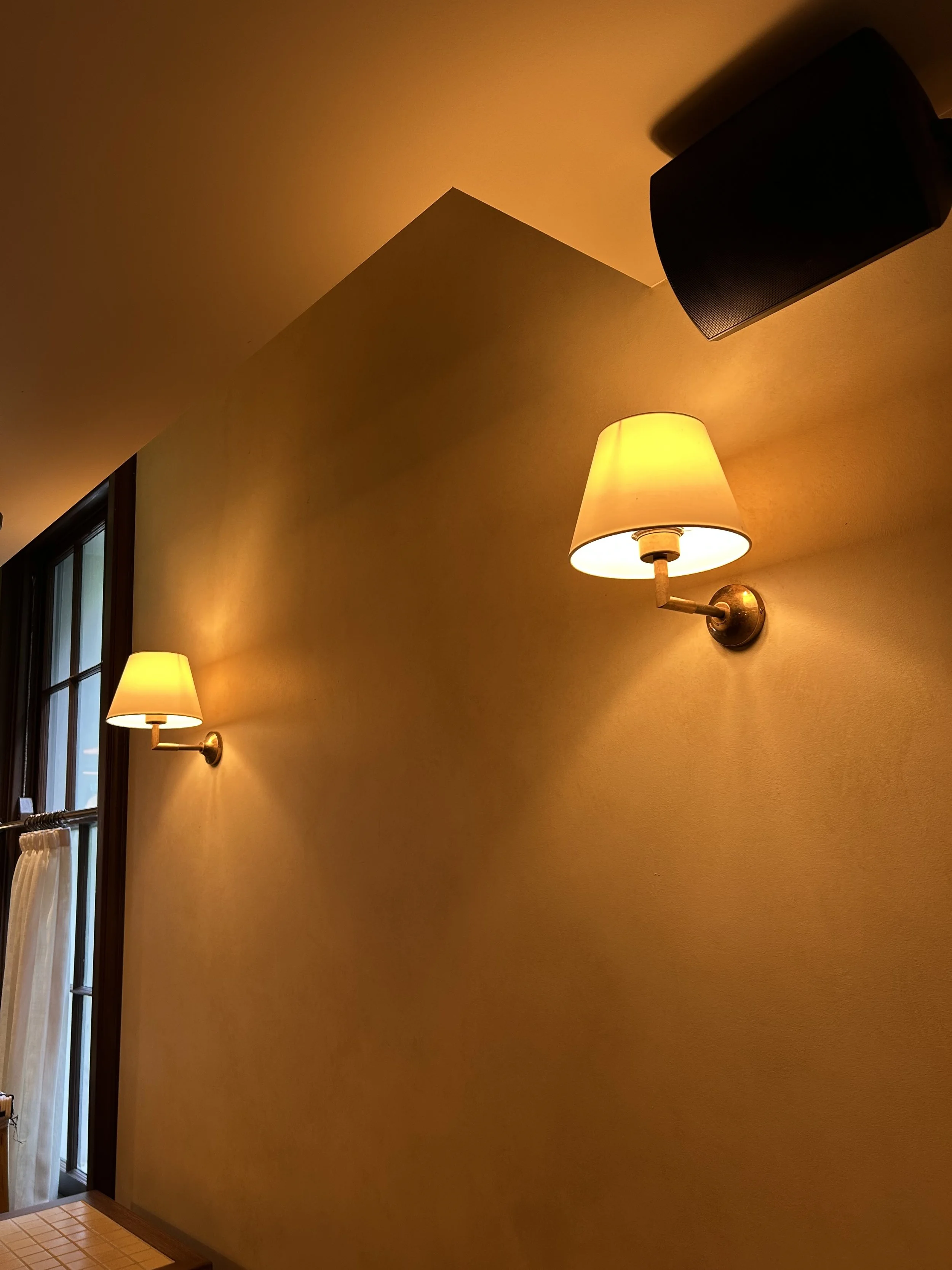 Two wall-mounted lamps with yellow shades illuminating a yellow wall in a room, adjacent to a large window with a curtain.
