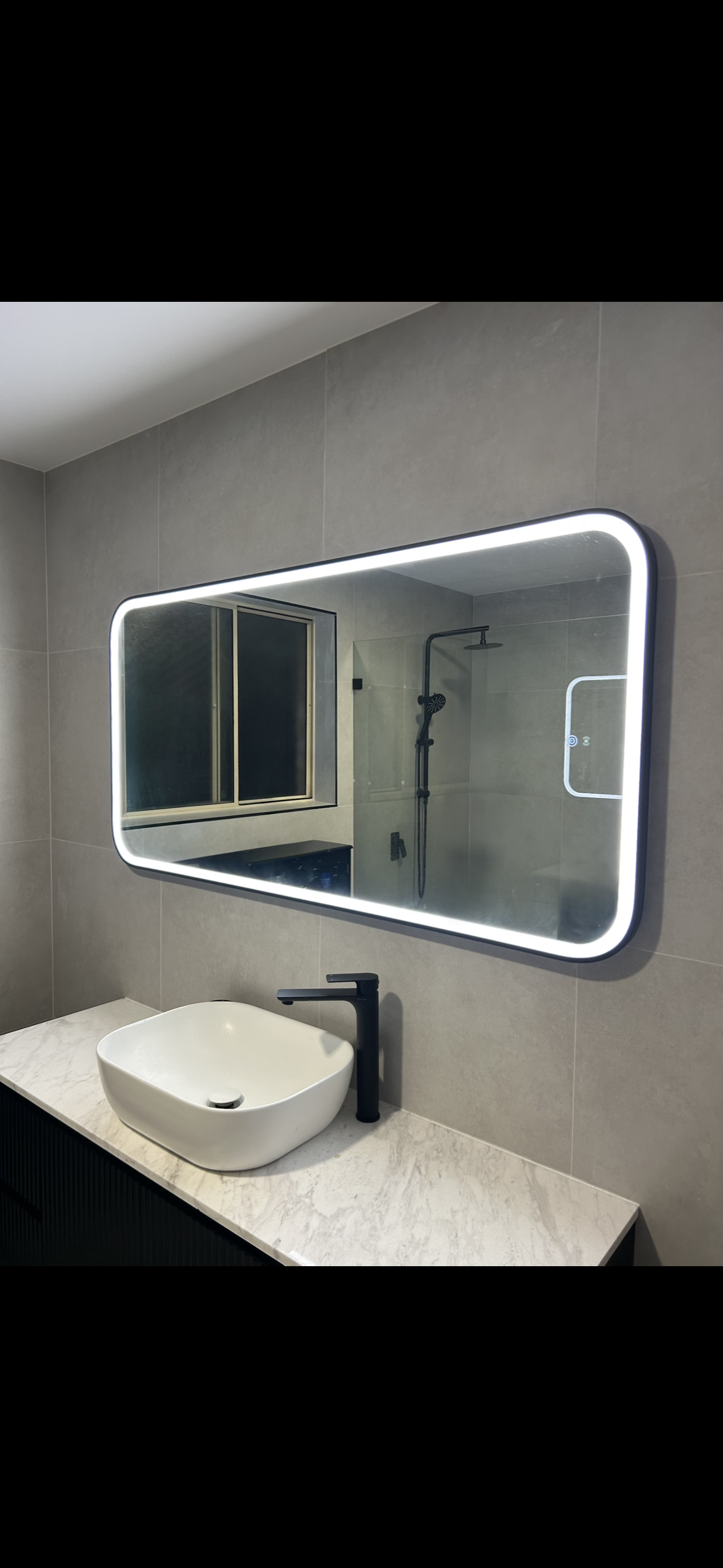 Modern bathroom with a large backlit mirror, white vessel sink, black faucet, gray tiled walls, and a shower area with a rain showerhead, seen through the mirror reflection.