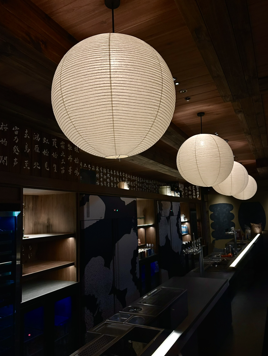 Interior of a bar or restaurant featuring hanging paper lanterns, dark wood paneling, and Japanese calligraphy artwork on the walls.