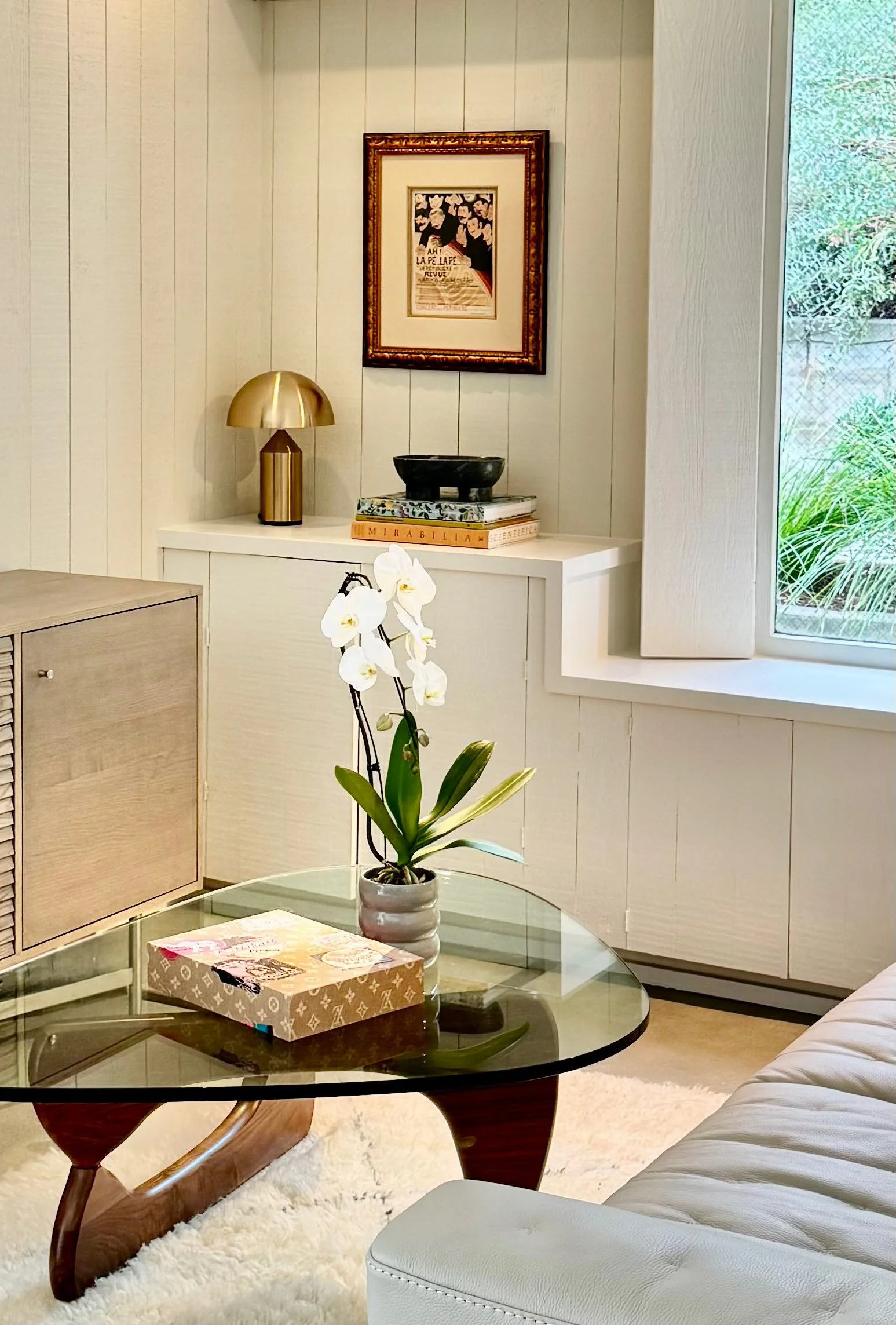 Living room with a glass coffee table, white sofa, potted orchid, framed artwork, decorative objects, and a window with outside greenery.