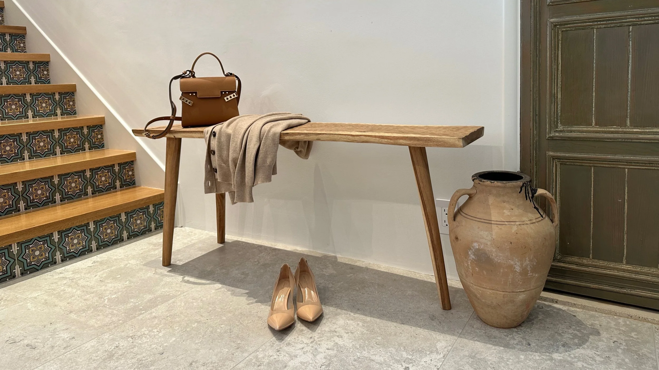 Wooden bench with a tan handbag and beige coat on top, beige high heels on the floor, a large ceramic vase beside the bench, a painted staircase to the left, and a wooden door to the right.