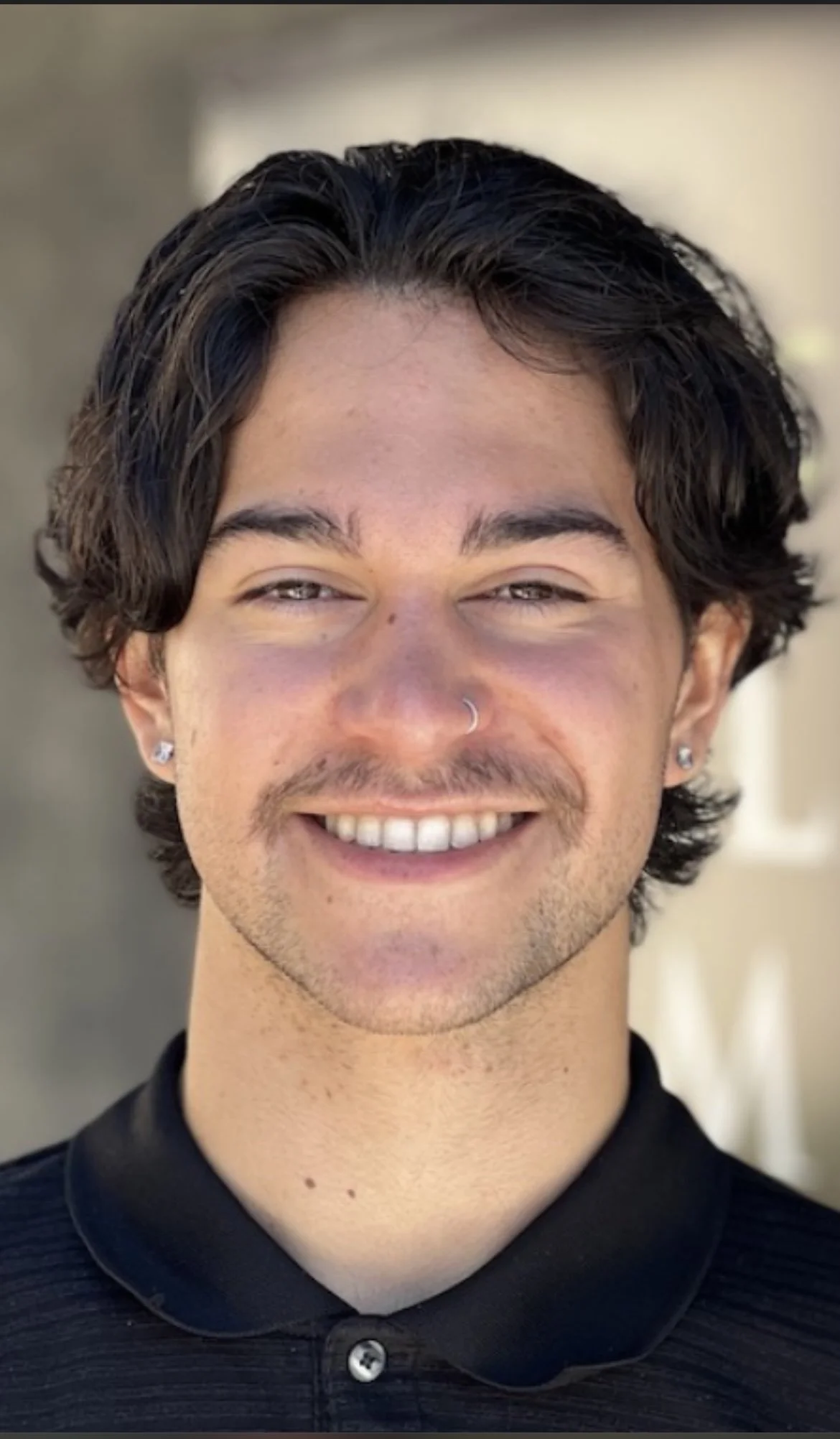 A young man with dark, wavy hair smiling, wearing earrings and a nose ring, dressed in a black collared shirt.