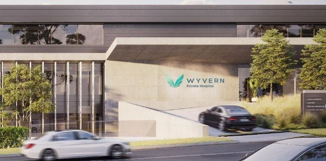 Exterior view of WYVERN Private Hospital building with cars driving past, some trees and greenery in front.
