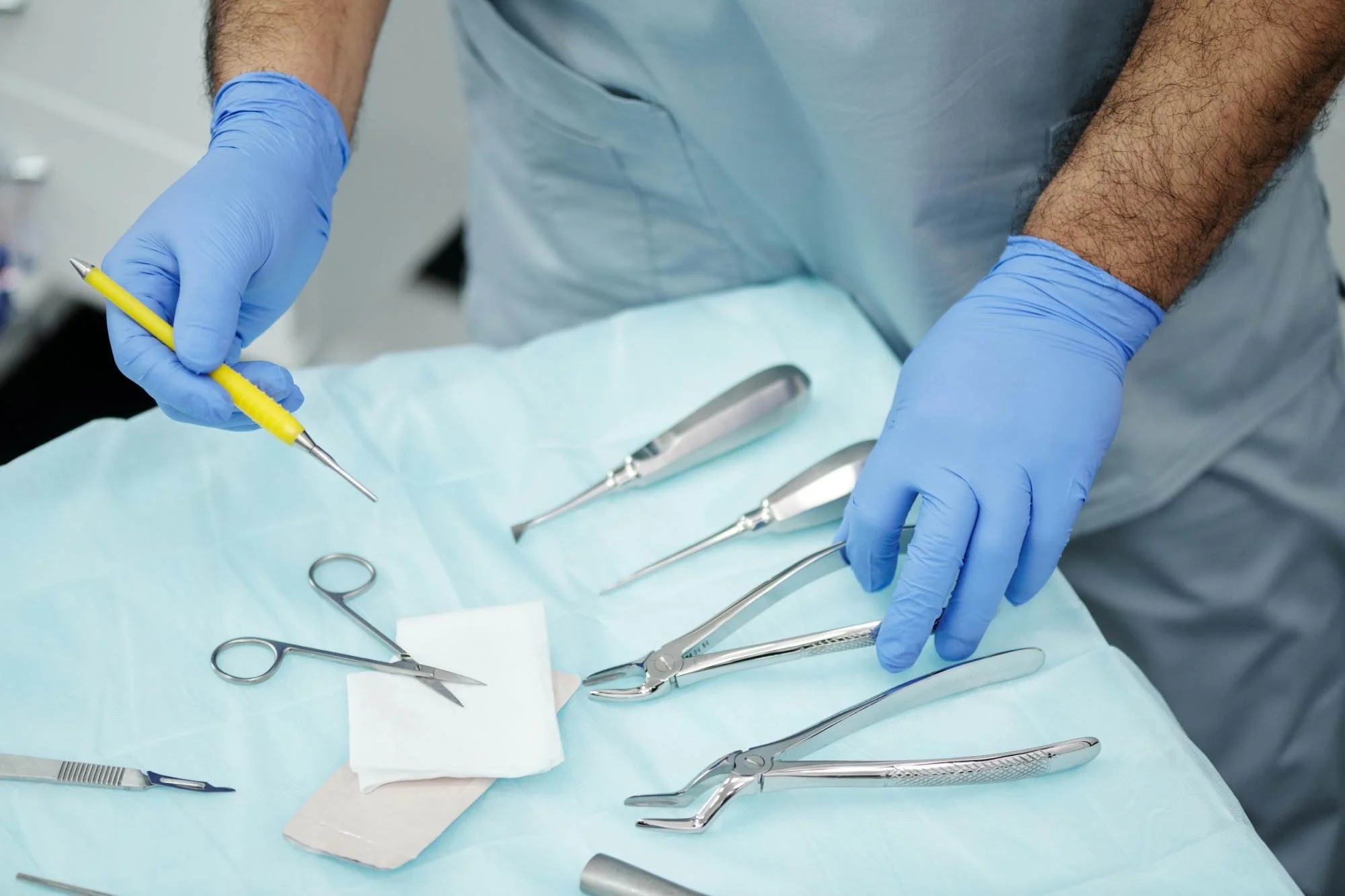 Close-up of a healthcare professional's hands wearing blue gloves, organizing surgical tools on a sterile drape, including scissors, forceps, and a scalpel.