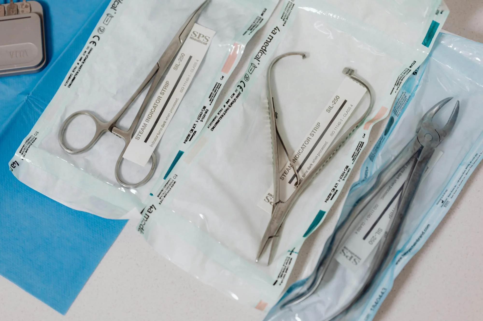 Sterile surgical instruments including forceps and surgical scissors in clear plastic packaging on a blue sterile drape, on a white countertop.