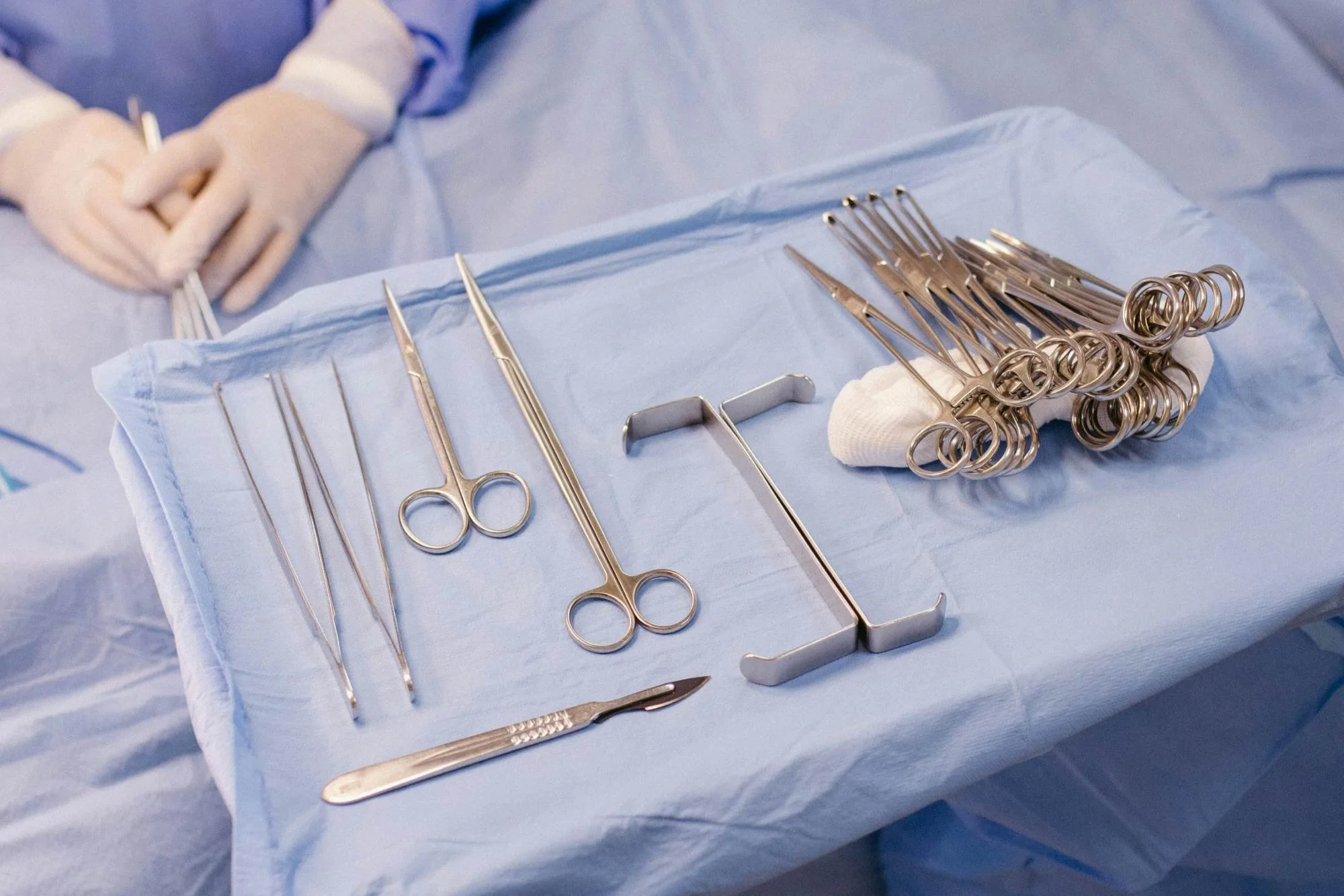 Silver surgical instruments arranged on a blue sterile drape, including scissors, forceps, and sutures, in a medical setting.