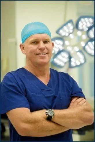 Dr Garett Smith surgeon wearing blue scrubs, a blue surgical cap, and a wristwatch, standing in an operating room with surgical lights in the background.