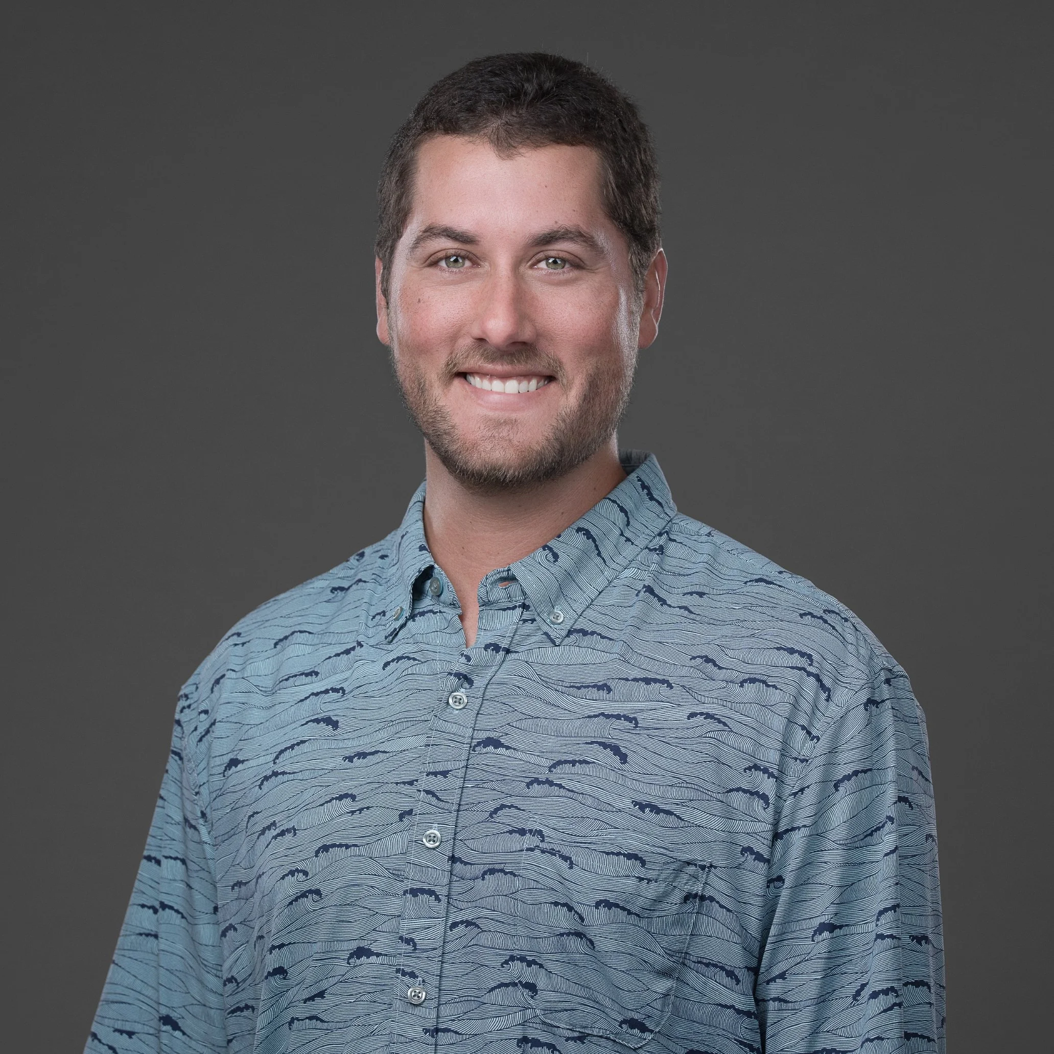 Headshot of a smiling man with short, dark hair and a light beard, wearing a patterned blue shirt, against a gray background.