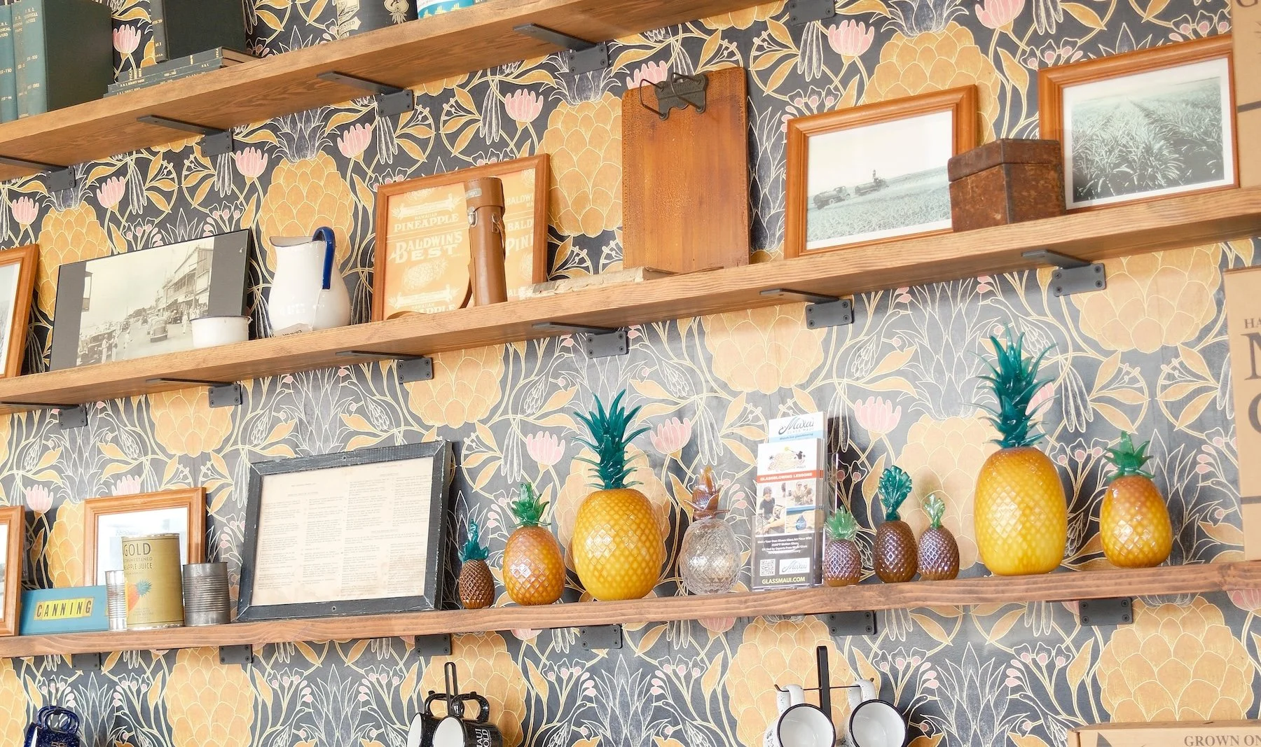 Decorative wall with three wooden shelves holding framed photos, vintage items, a can of pineapple juice, pineapples in various sizes, and a patterned wallpaper background.