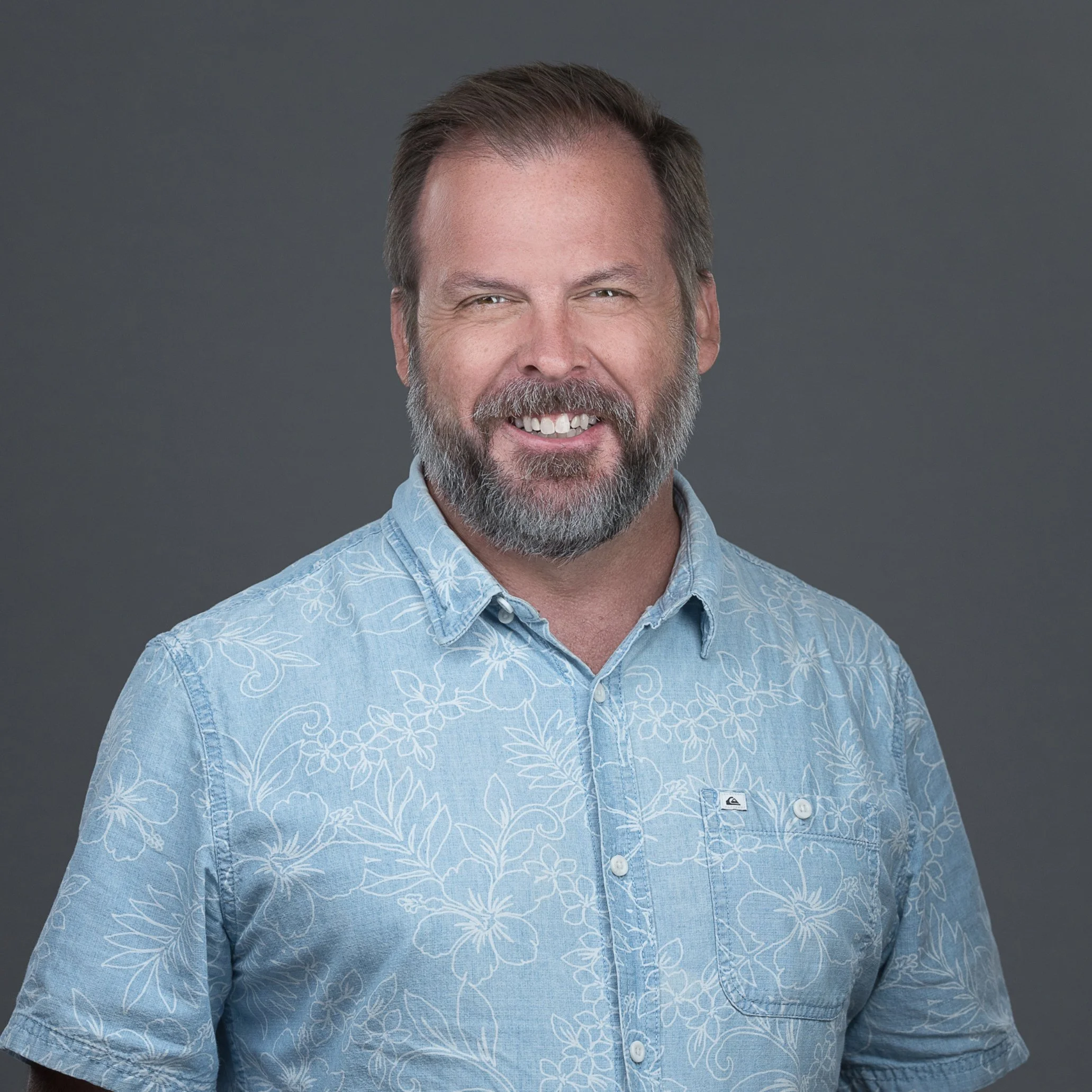 Close-up of a middle-aged man with a beard and short brown hair smiling, wearing a light blue Hawaiian shirt with white floral patterns, against a plain gray background.