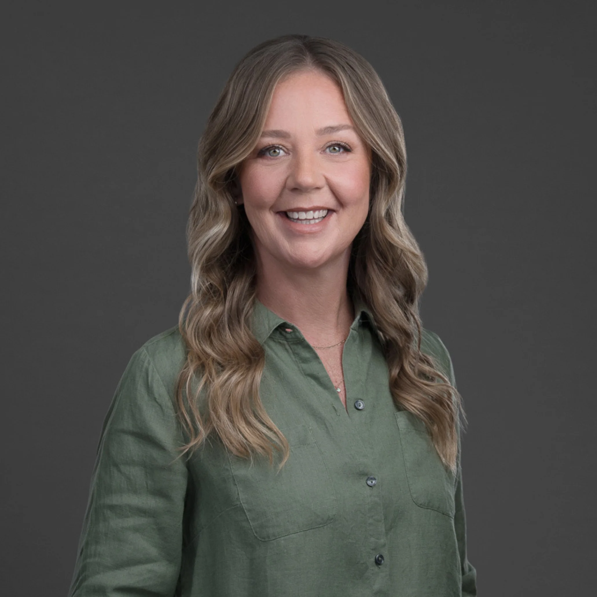 Close-up of a smiling woman with long wavy blonde hair, wearing a green button-up shirt, against a gray background.