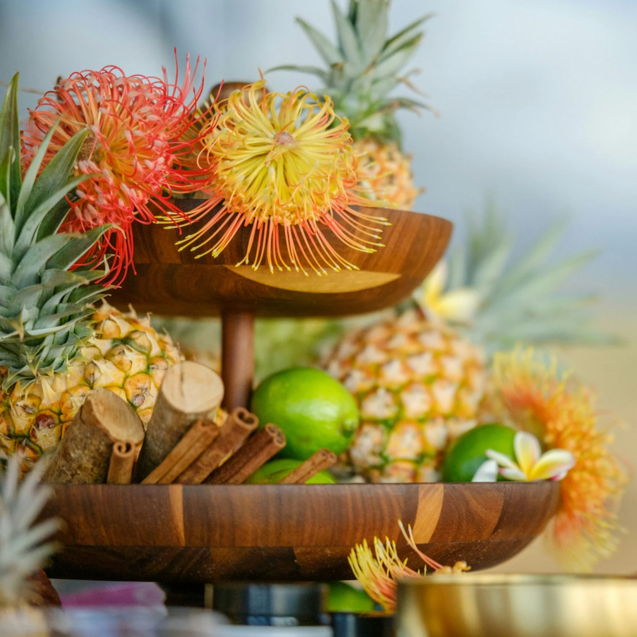 Close-up of a decorative arrangement of pineapples, green apples, cinnamon sticks, and tropical flowers in a wooden bowl.