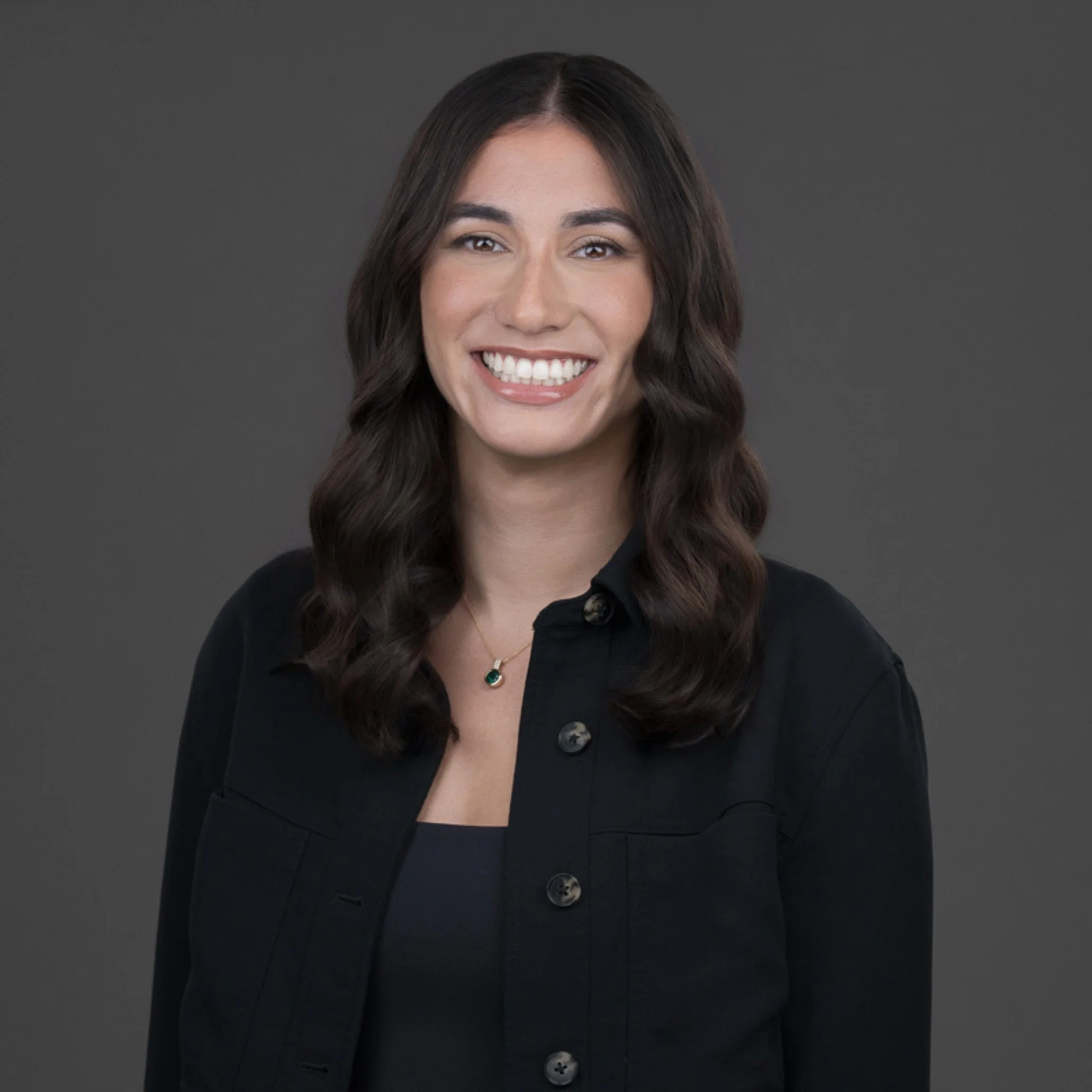 A woman with long dark hair, smiling, wearing a black collared shirt and a necklace with a green pendant, standing against a gray background.