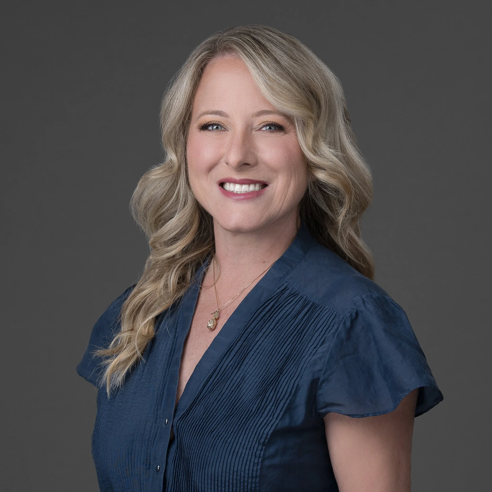 Close-up photo of a middle-aged woman with wavy blonde hair, blue eyes, smiling showing her teeth, wearing a dark blue blazer and a necklace with a pendant, against a gray background.
