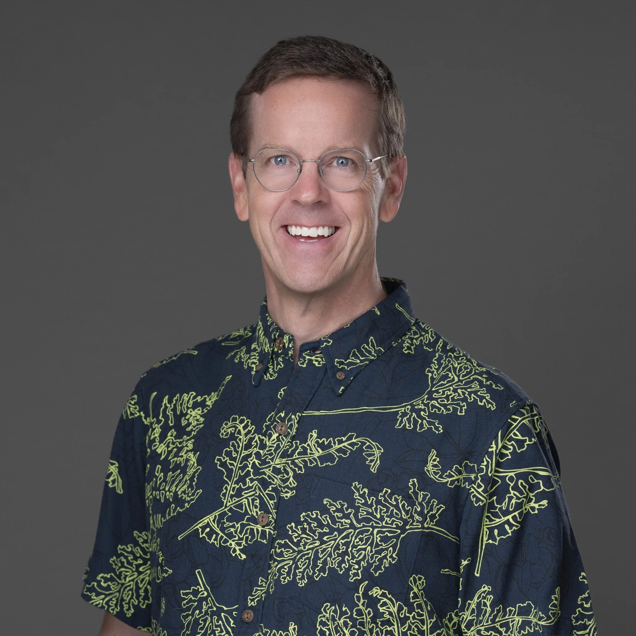 A smiling man with short brown hair, wearing round glasses and a navy shirt with a yellow line pattern resembling interconnected leaves or branches, standing against a gray background.