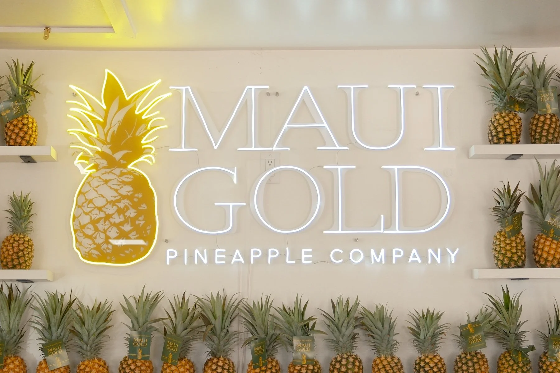 Neon sign for Maui Gold Pineapple Company with a large illuminated pineapple graphic, surrounded by shelves of fresh pineapples with Maui Gold tags.