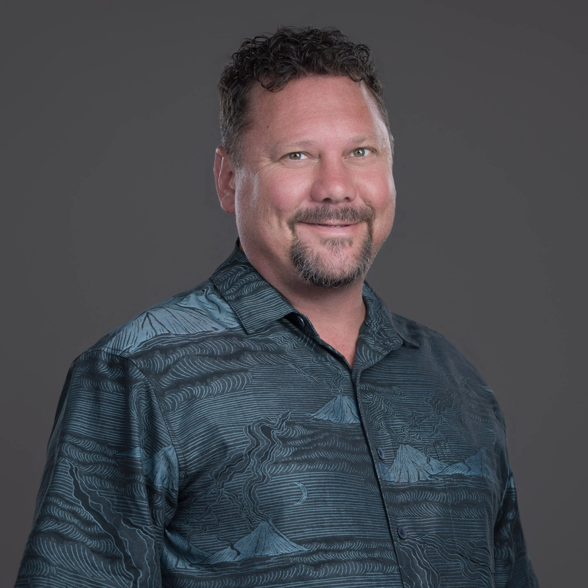 Headshot of a middle-aged man with curly dark hair, light skin, green eyes, a goatee, wearing a dark patterned shirt, smiling against a plain gray background.