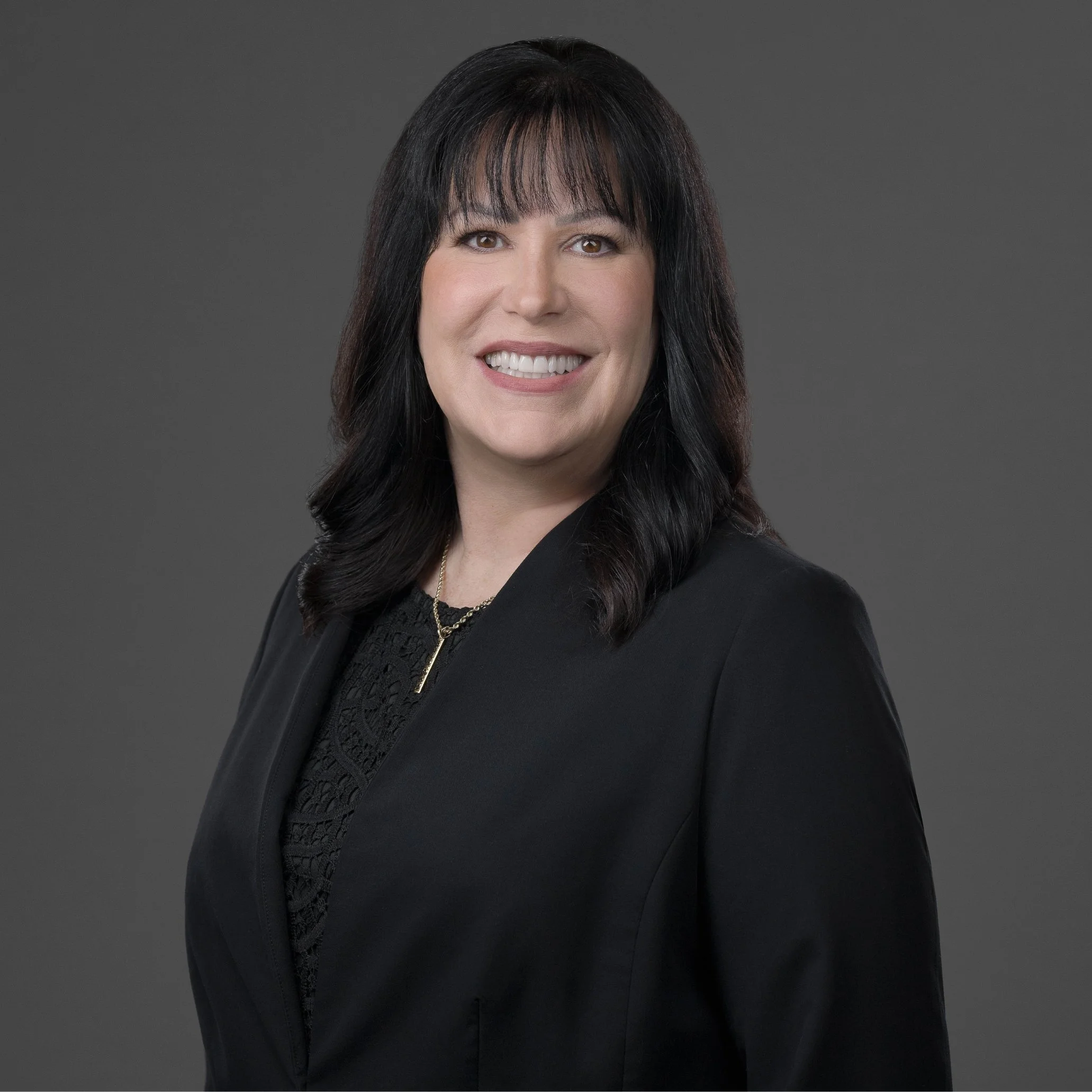 A woman with dark hair, light skin, and a smile wearing a black blazer over a patterned black top, standing against a dark gray background.